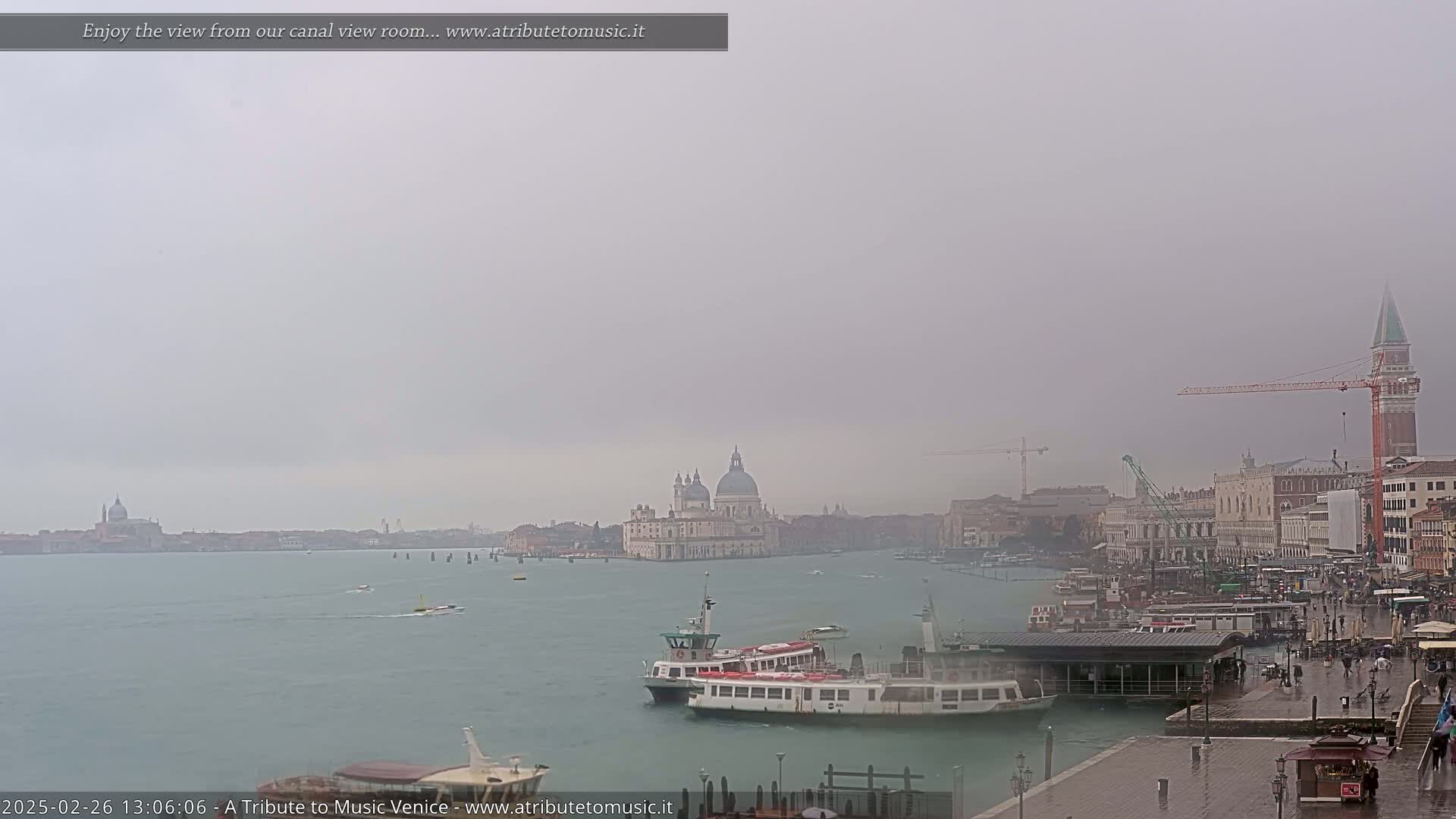 A hazy, rainy view of Venice shows several boats on a canal with the Basilica di Santa Maria della Salute and other buildings in the distance.