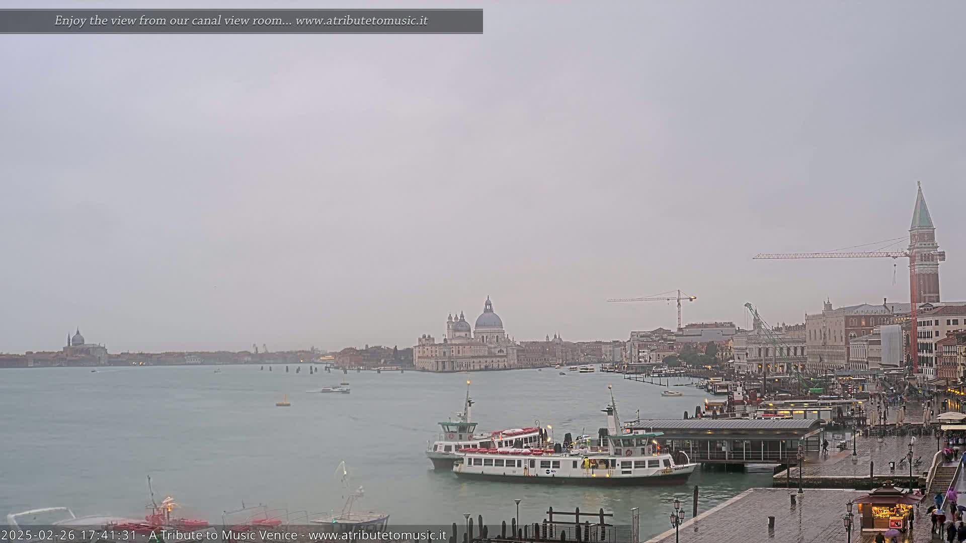 A hazy, overcast day shows a Venetian canal scene with several boats, including passenger ferries, moored near a waterfront building, with the Basilica di Santa Maria della Salute and the bell tower of St. Mark's Square visible in the distance.
