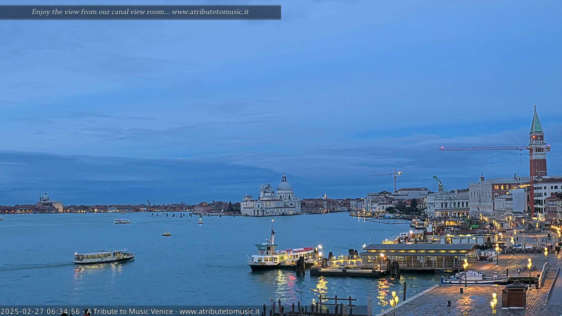 Under a twilight blue sky, a calm body of water shows several boats near a waterfront city, with large buildings and a dome-topped structure visible in the distance.