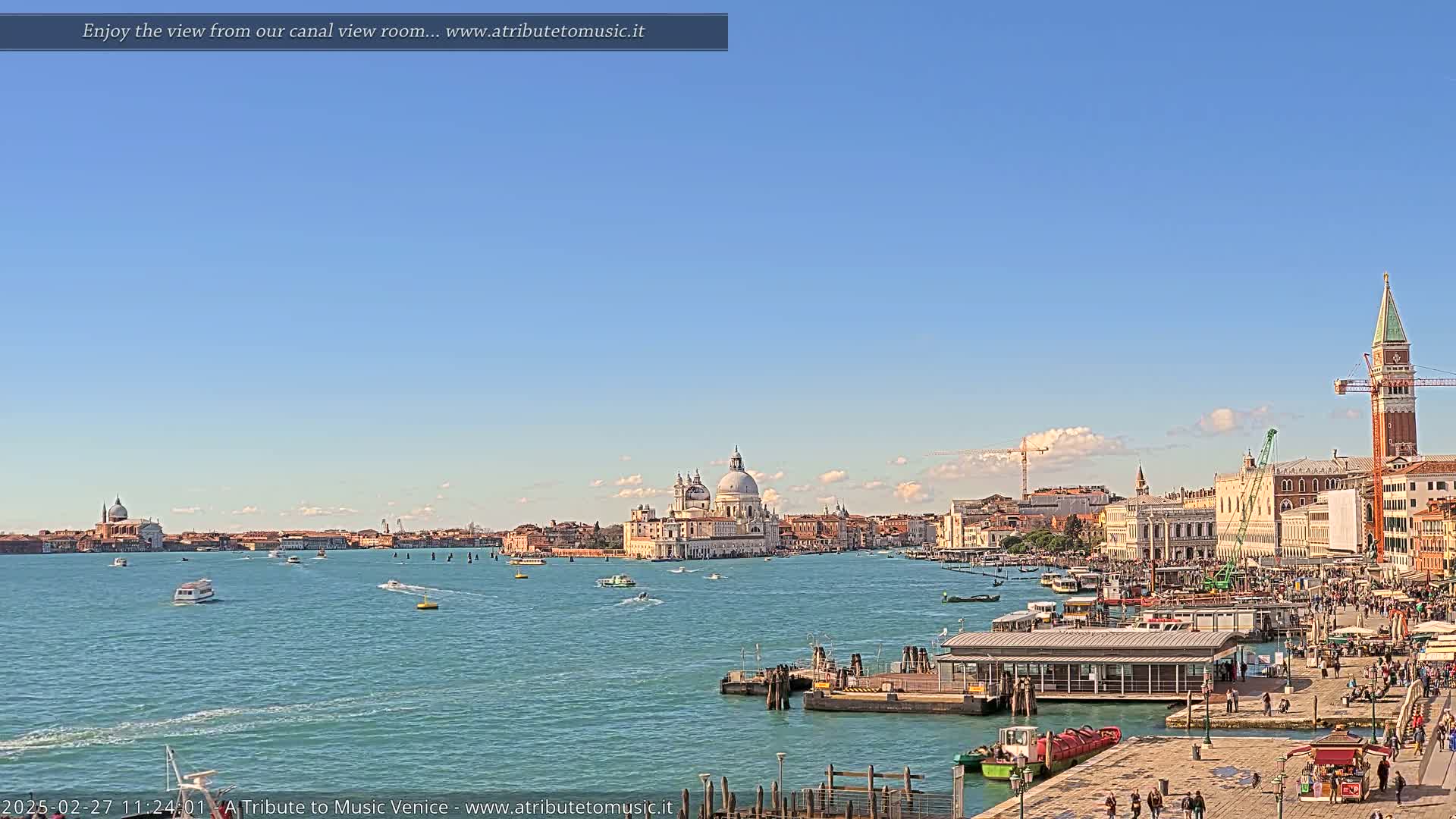 A wide shot of a Venetian canal on a clear, sunny day shows a large body of water with boats, buildings, and a church in the background.