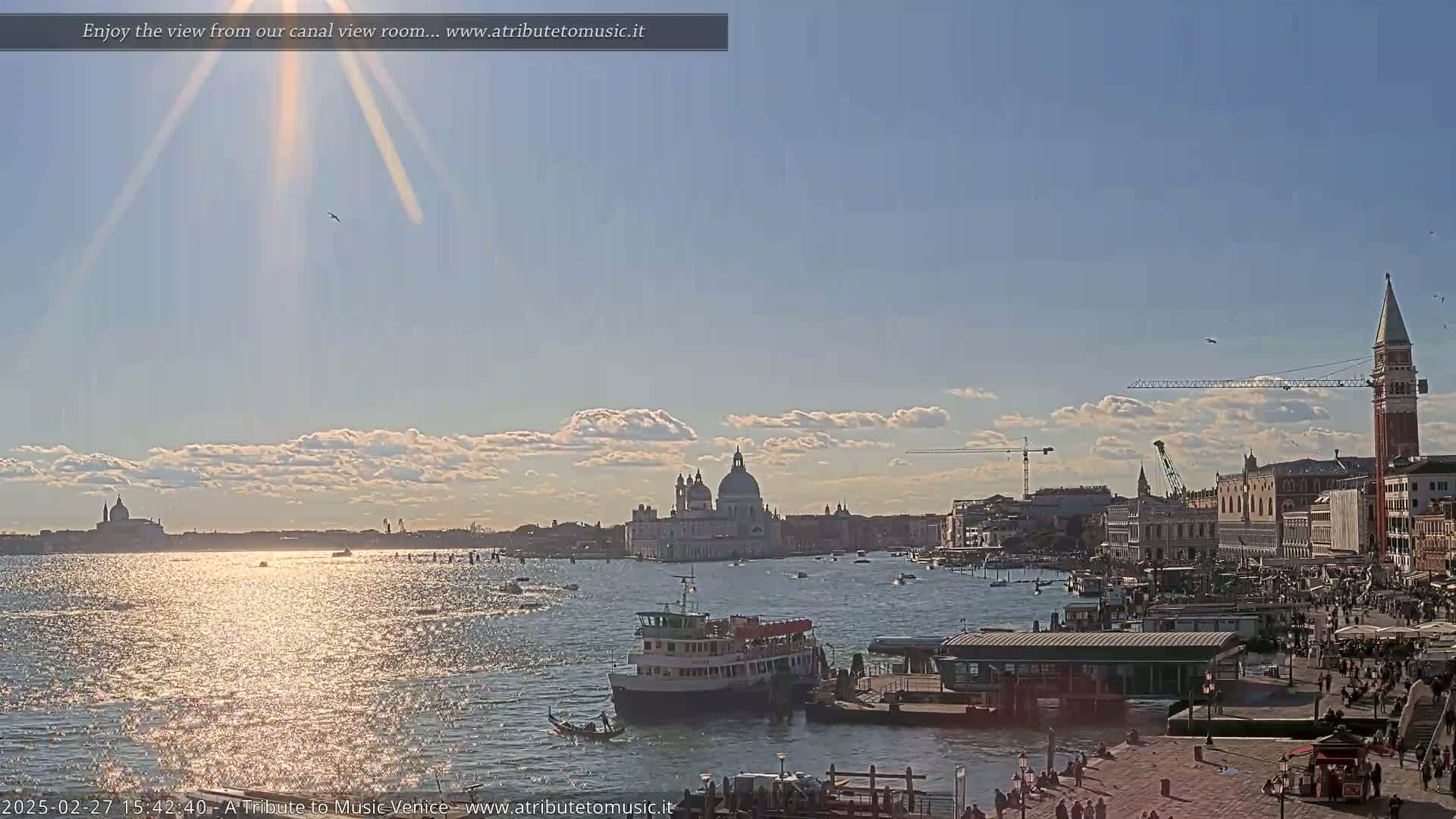 A sunny day in Venice shows a wide canal with a ferry, a waterfront with people, and the Basilica di Santa Maria della Salute in the distance.