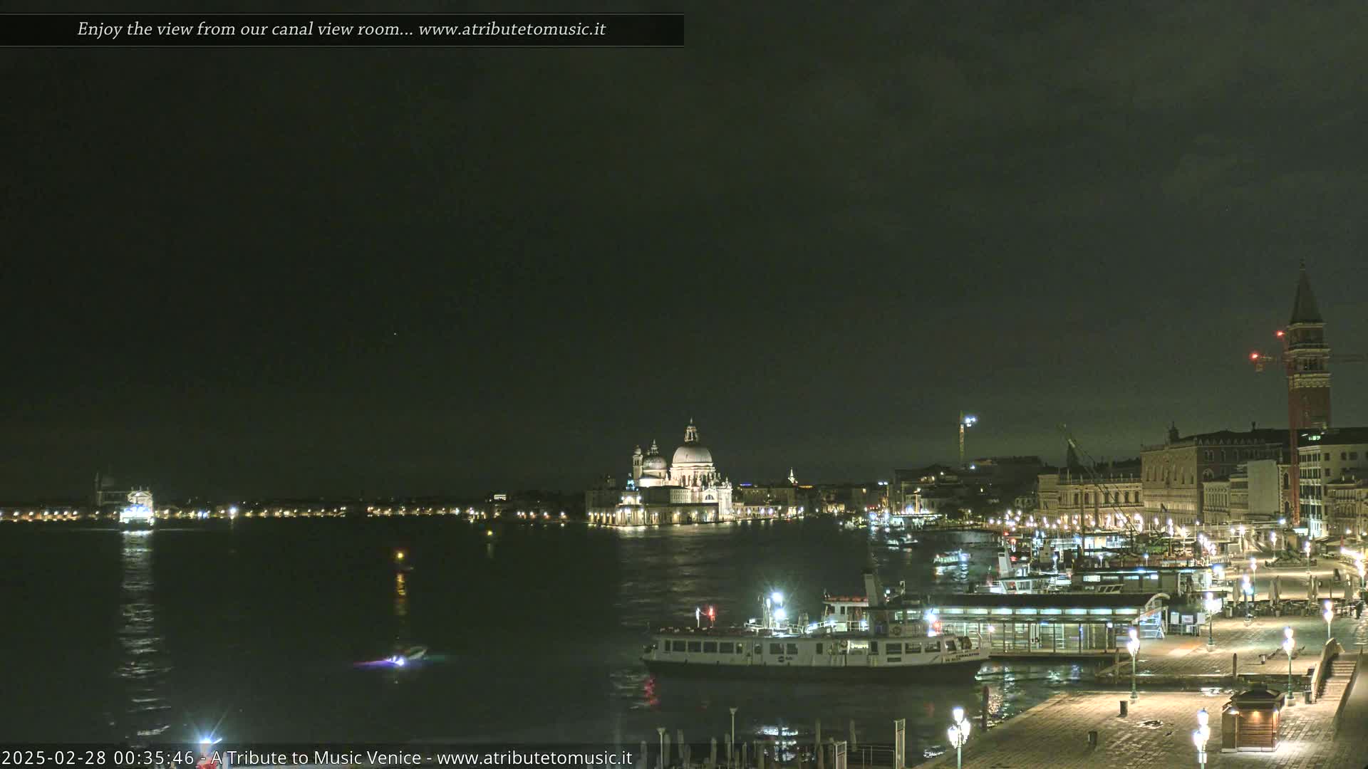 A nighttime view of a Venetian canal shows boats moored along a waterfront, with the Basilica di Santa Maria della Salute and other buildings illuminated across the water under a dark, cloudy sky.