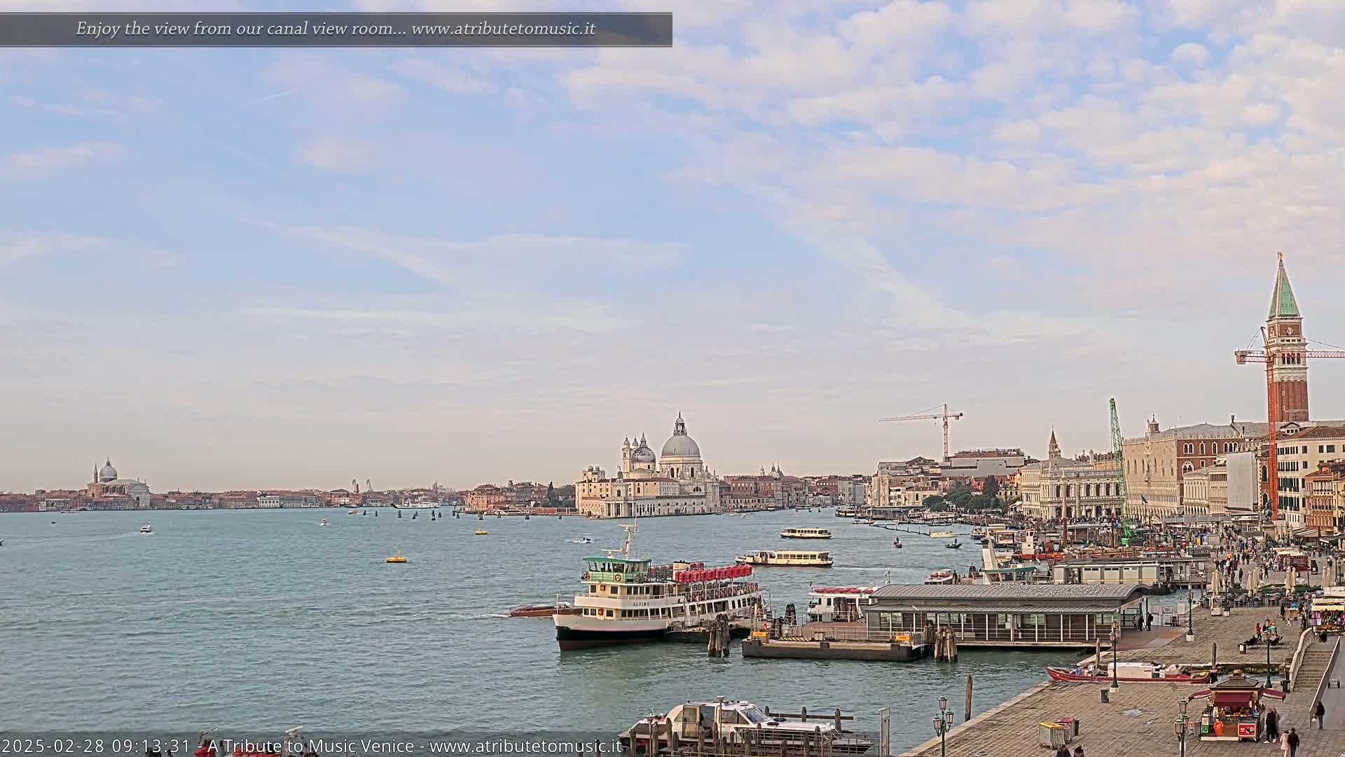 A wide view of a waterway in Venice, Italy, on a partly cloudy day, shows several water taxis and boats, with the Basilica di Santa Maria della Salute and St. Mark's Campanile visible in the distance.