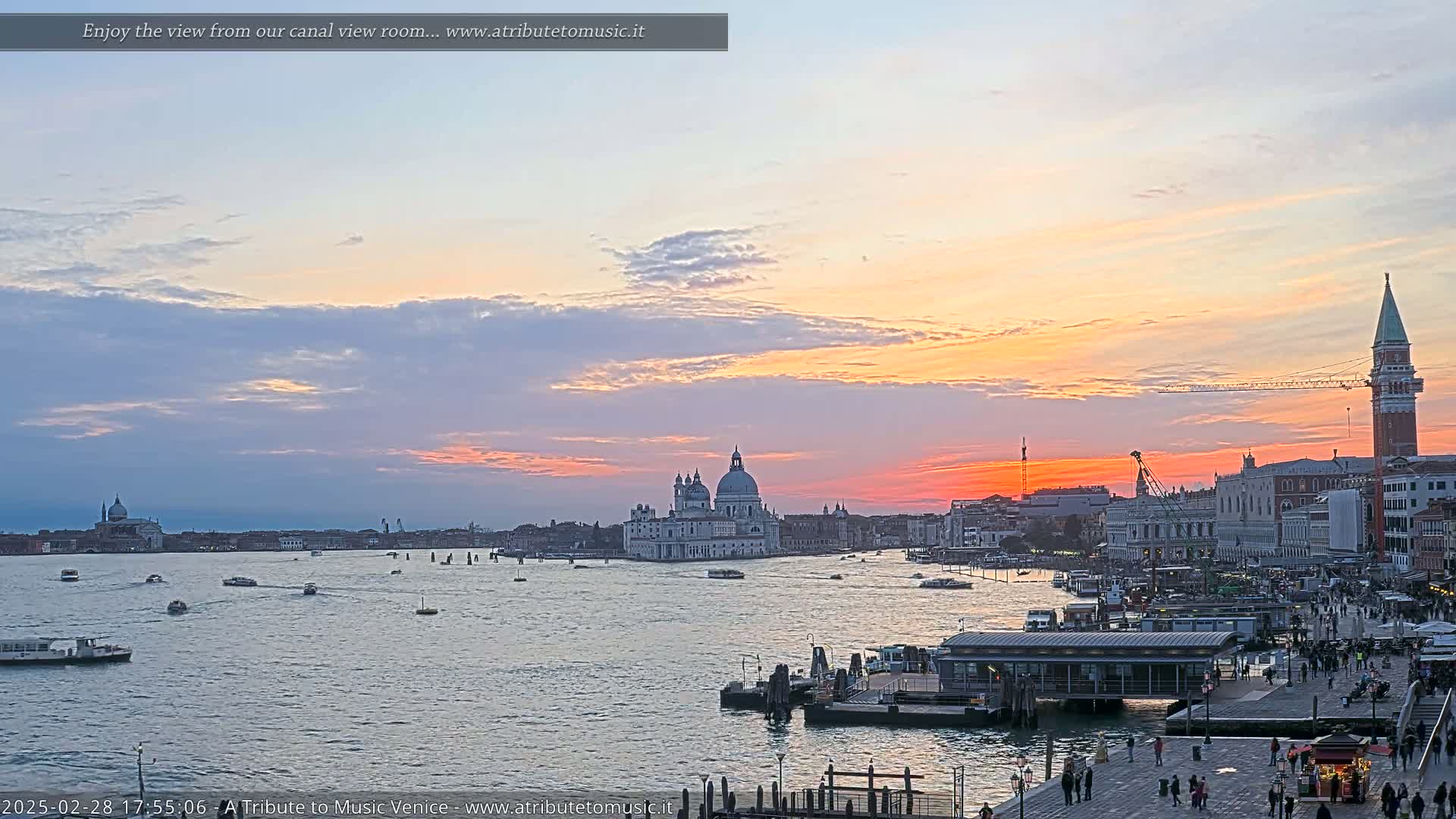 A Venetian sunset over a calm canal, showing boats, buildings, and a large church, with a partly cloudy sky displaying warm orange and pink hues.