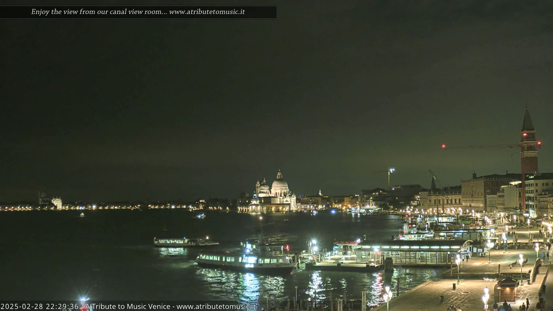 A nighttime view of a canal in Venice, Italy, shows several boats and illuminated buildings along the waterfront under a dark sky.