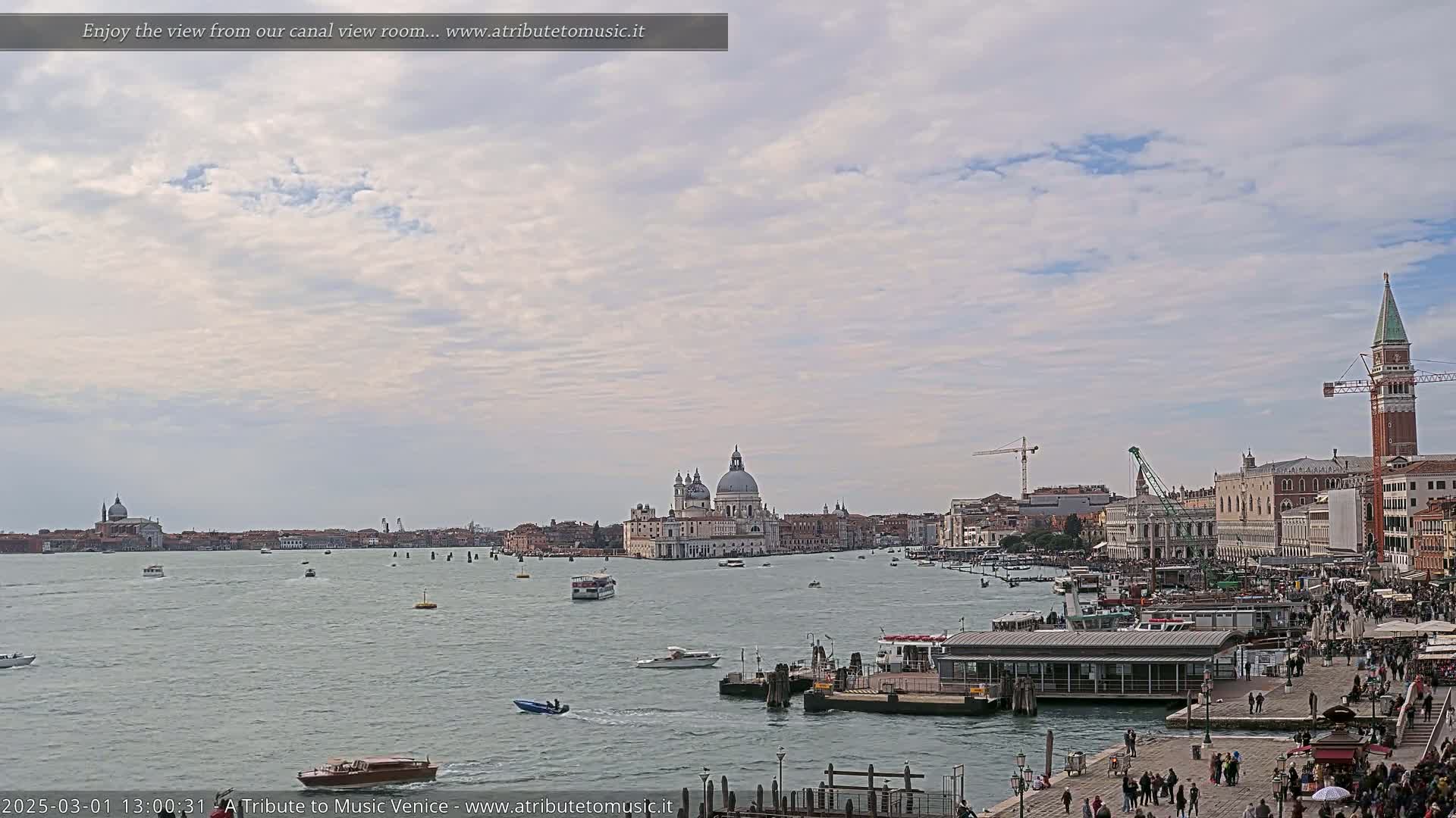 A wide, calm canal in Venice, under a partly cloudy sky, shows numerous boats and buildings, including a large domed church in the distance.