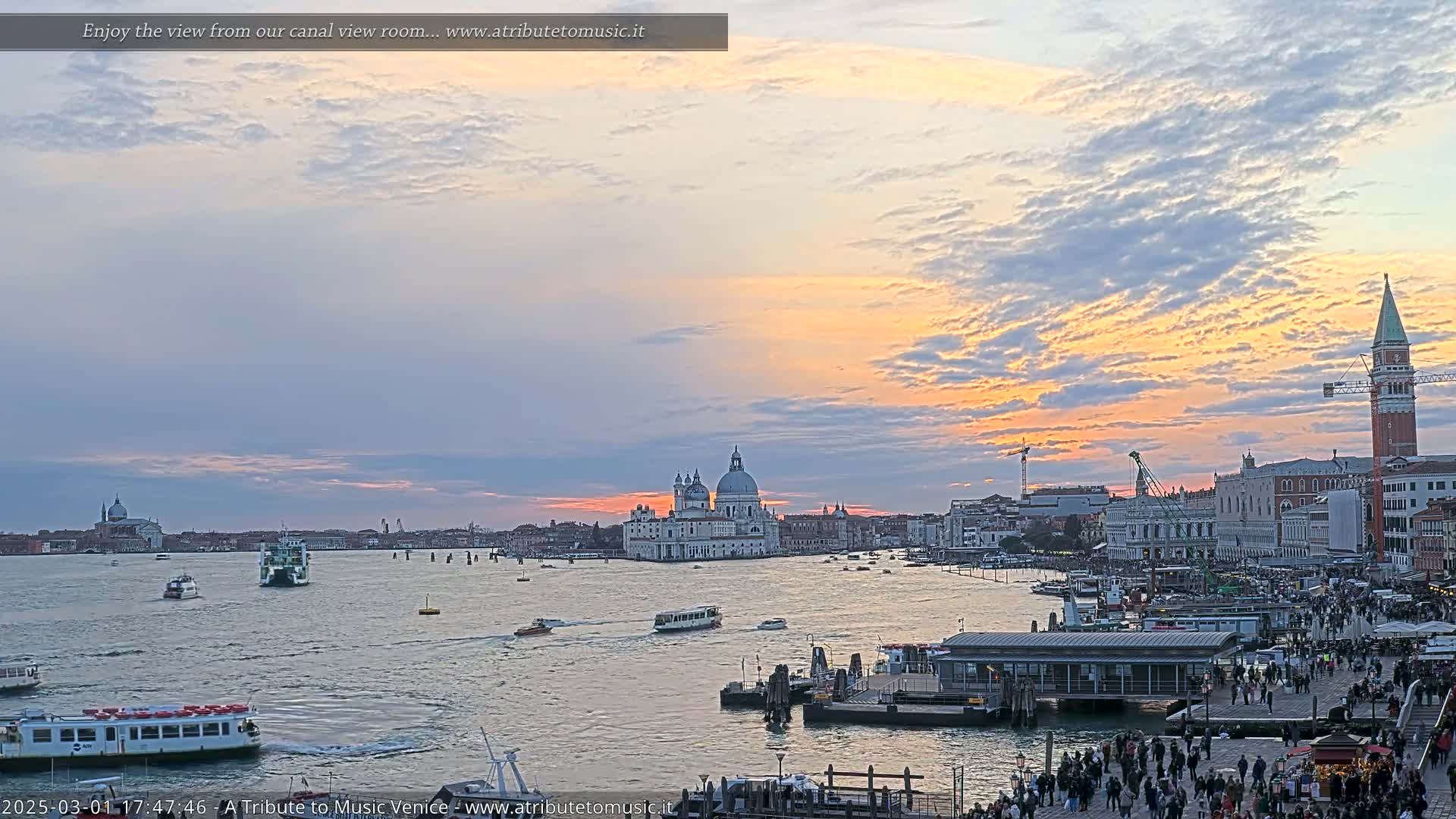 A sunset over Venice shows a calm canal with boats and many people gathered near a waterfront building, with the Basilica di Santa Maria della Salute prominent in the background.