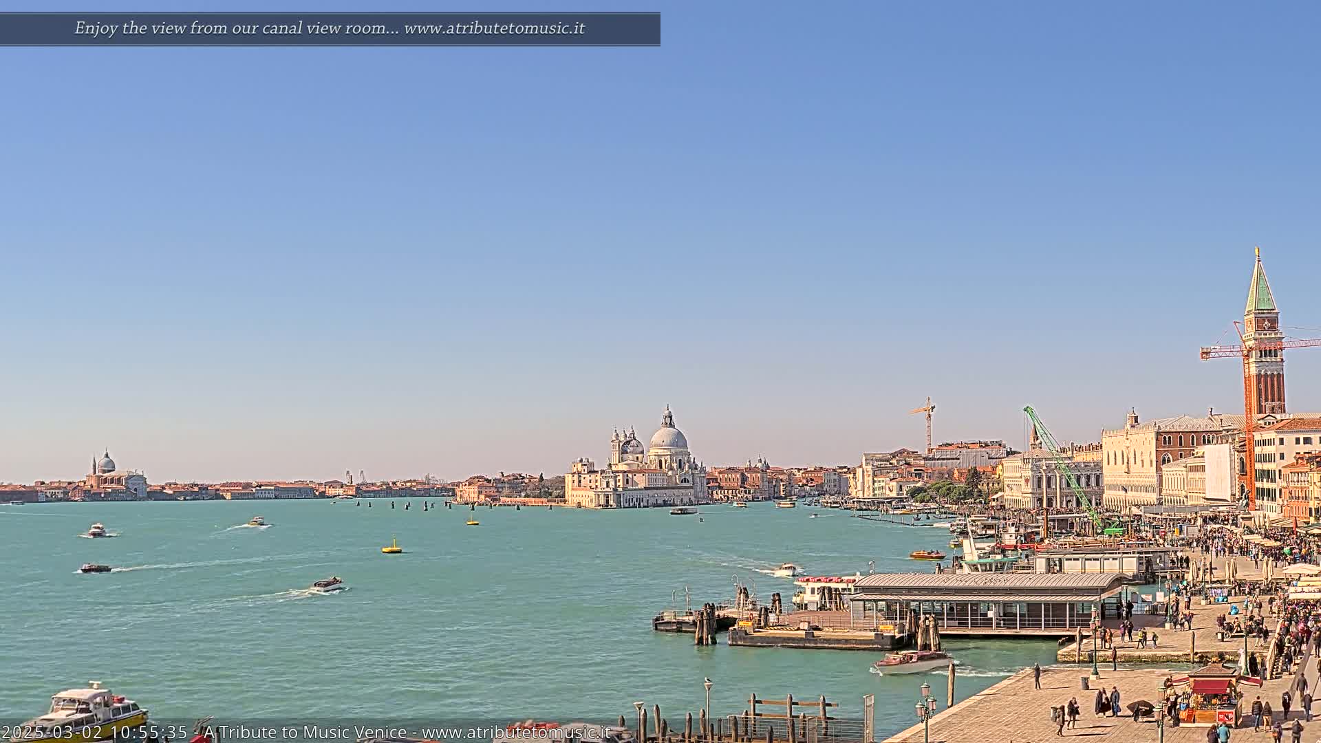 A sunny day in Venice shows a wide canal with boats, a waterfront promenade with pedestrians, and the Basilica di Santa Maria della Salute in the distance.