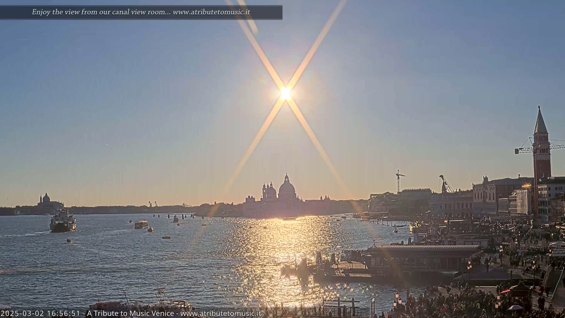 A bright, sunny day over Venice shows a canal with boats and people on the waterfront, the Basilica di Santa Maria della Salute in the distance.