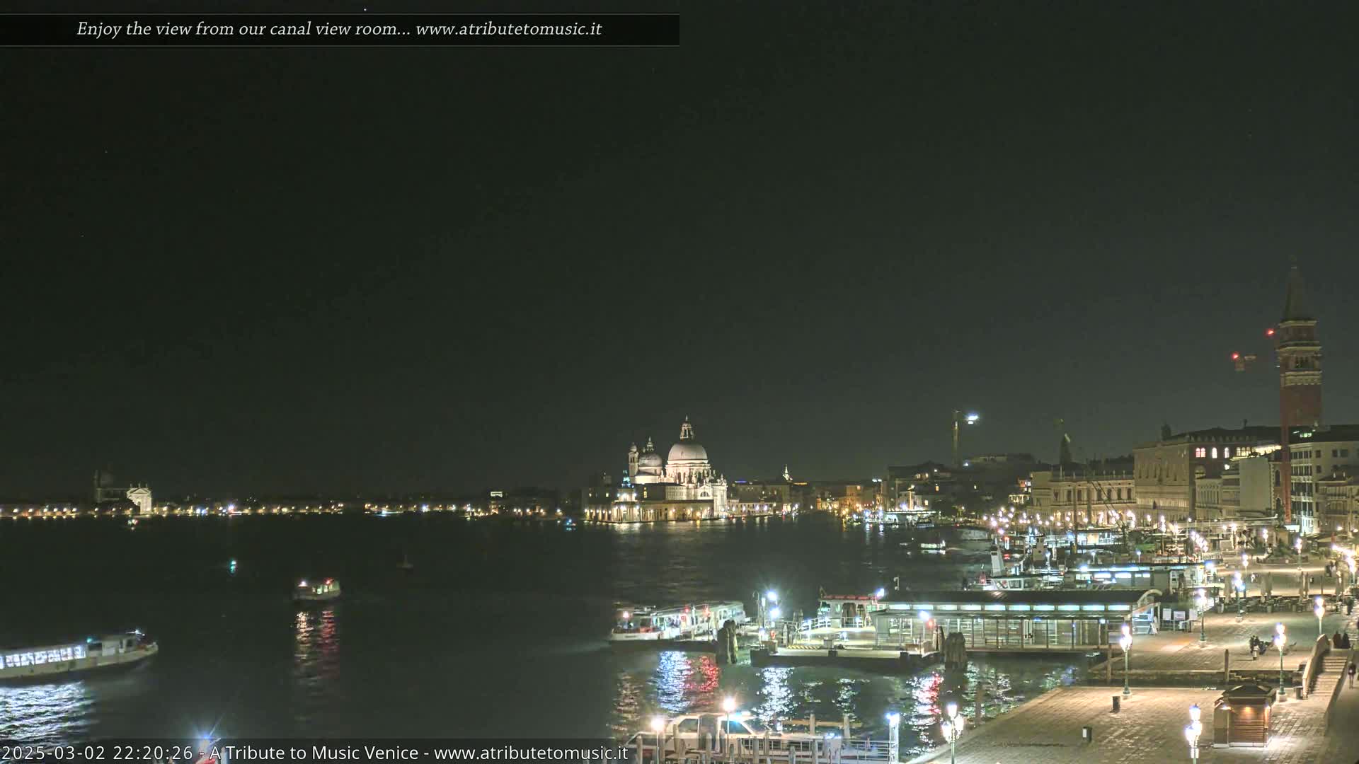 A nighttime view of a canal in Venice, Italy, shows boats on the water and illuminated buildings along the shore, under a clear, dark sky.