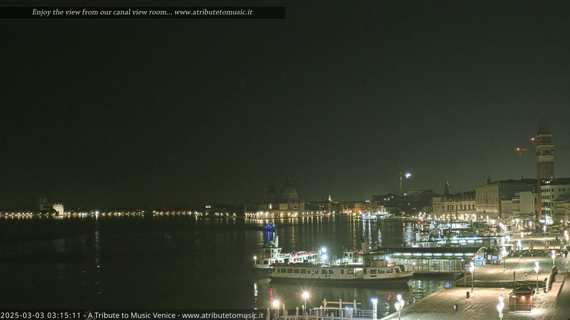 A nighttime view across a calm canal shows illuminated buildings and a few boats docked at a waterfront.