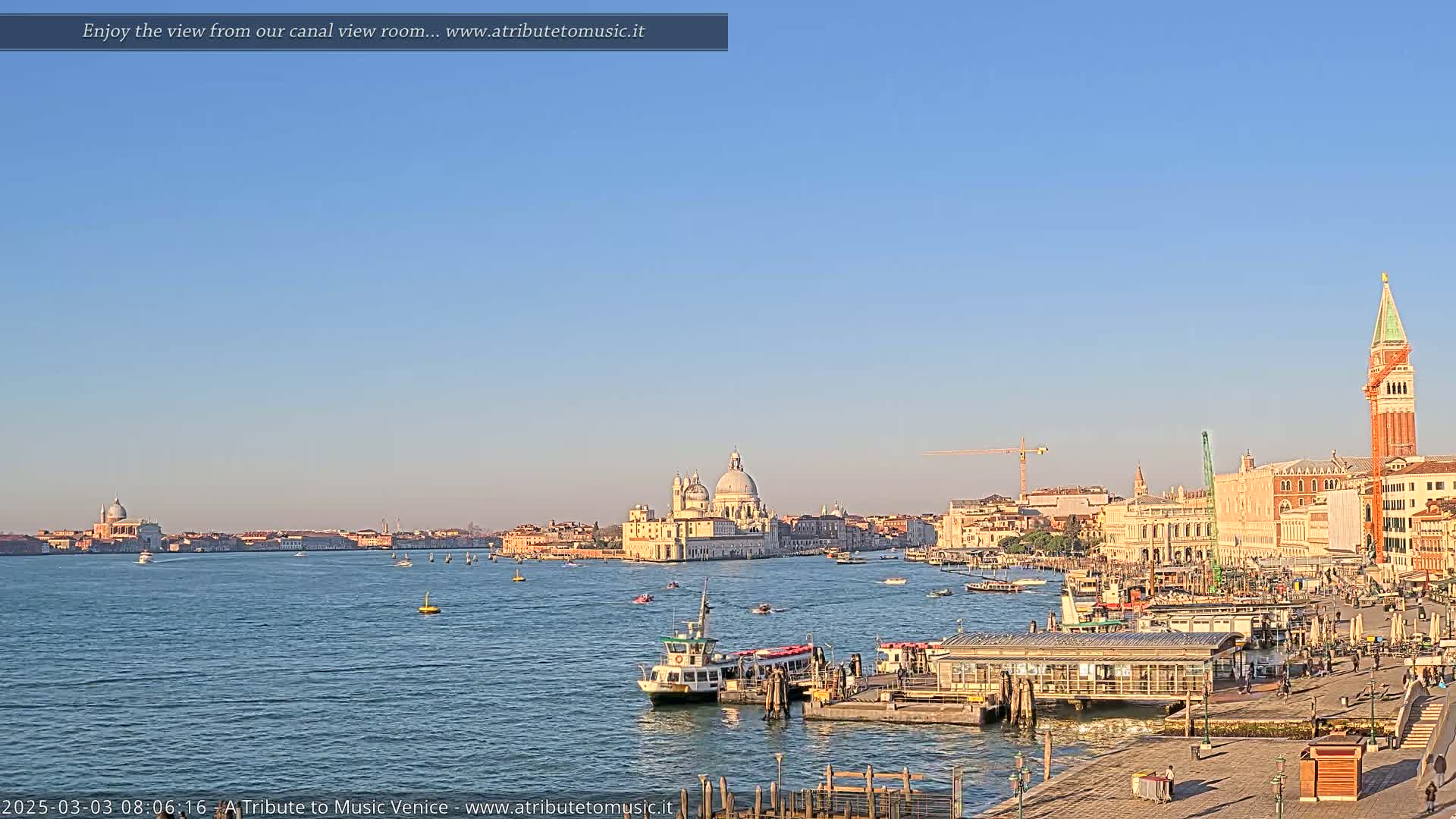 A clear, sunny day in Venice shows a wide canal with boats, a waterfront with buildings, and the iconic domes of a church in the distance.