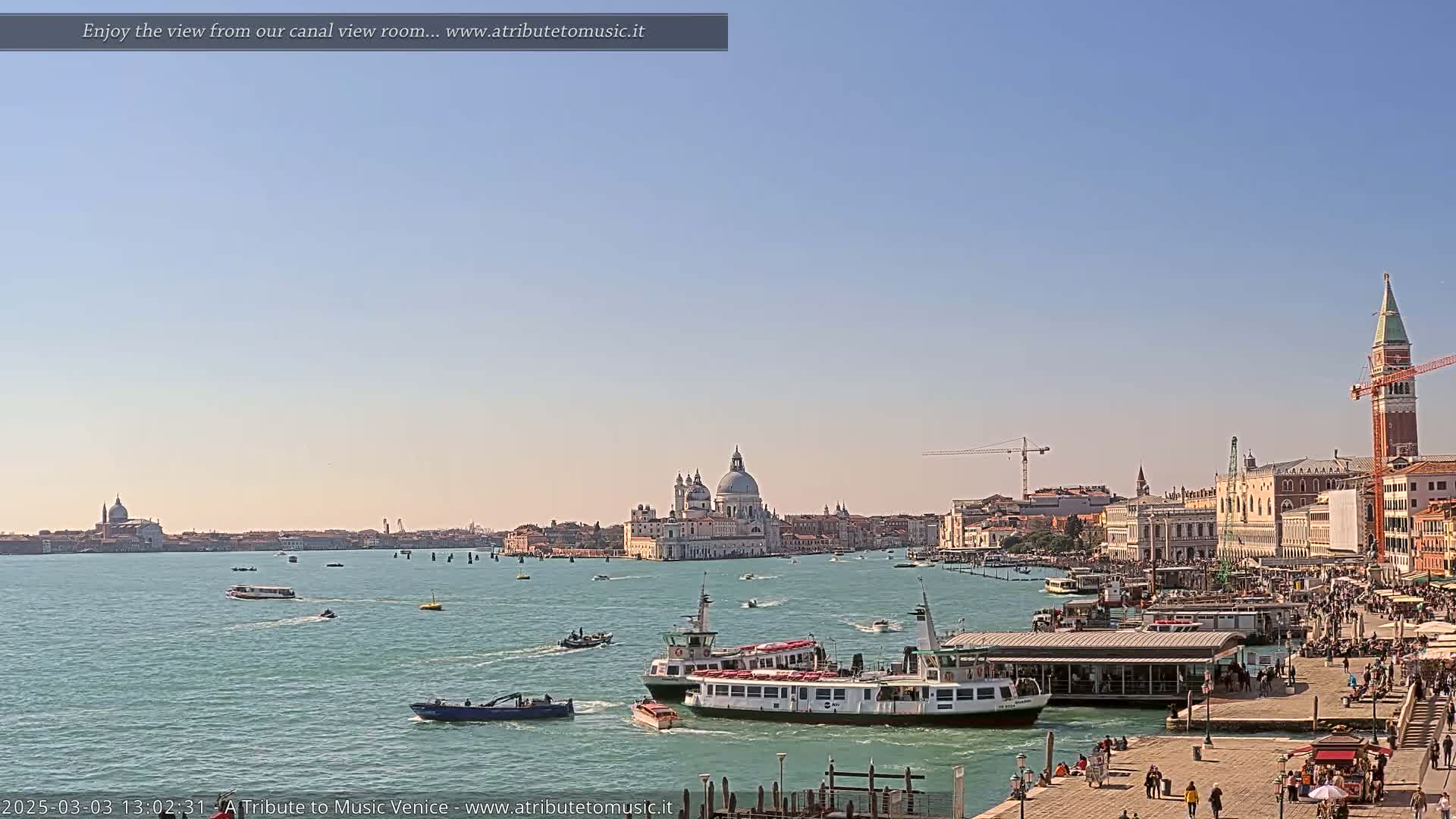 A sunny day in Venice shows a wide canal with several boats, a waterfront bustling with people, and the iconic Basilica di Santa Maria della Salute in the distance.