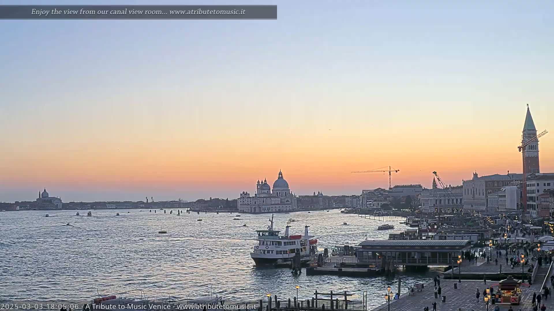 A Venetian canal scene at sunset shows a ferry docked near a waterfront promenade, with the Basilica di Santa Maria della Salute and San Giorgio Maggiore in the distance.