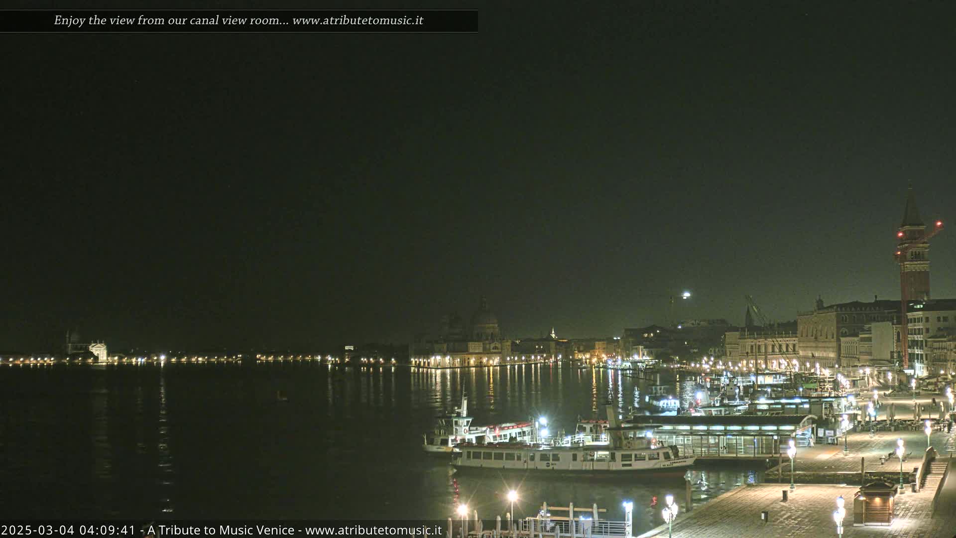 A nighttime view across a calm canal shows illuminated buildings, boats, and a waterfront walkway under a dark, clear sky.