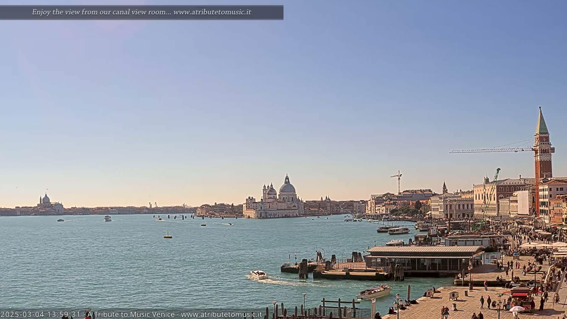A sunny day in Venice shows a wide canal with boats and buildings along the shore, including a large domed church, and a tall bell tower.