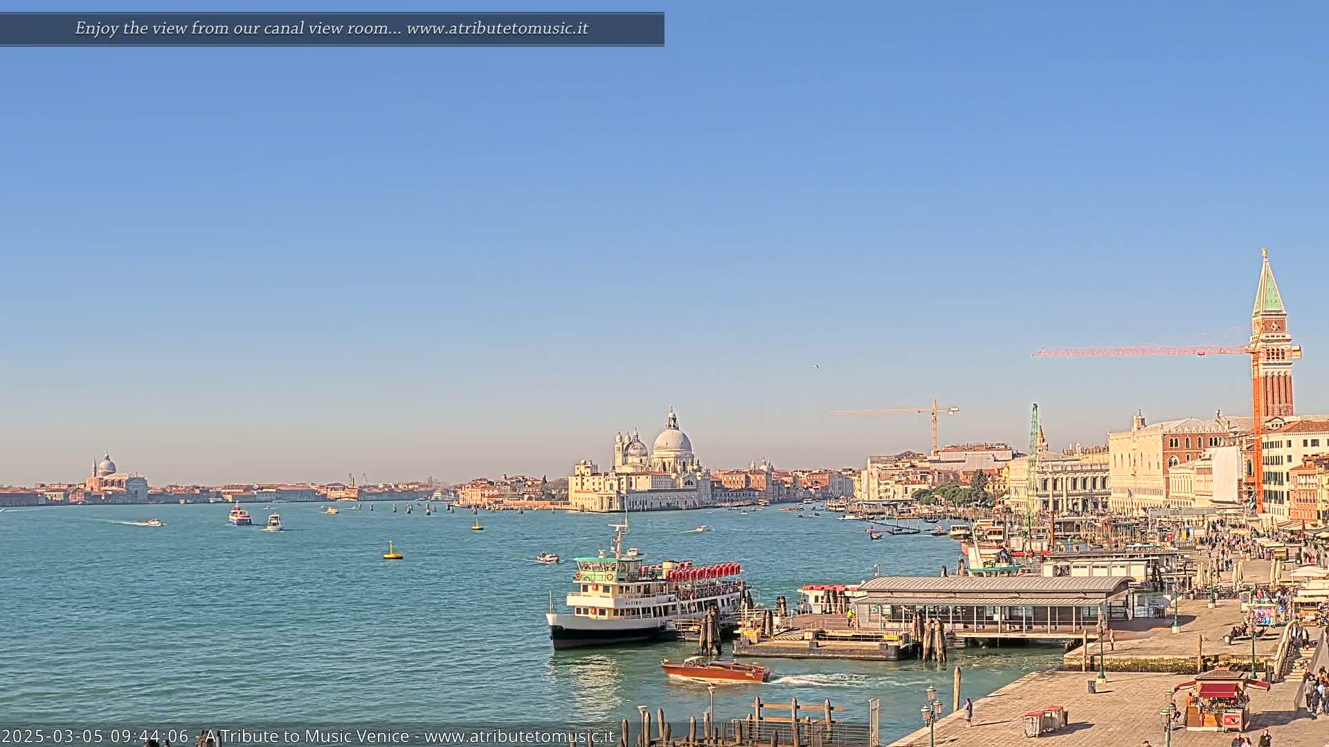 A sunny day in Venice shows a wide canal with several boats and a view of the city skyline, including a large domed church and the San Giorgio Maggiore bell tower.