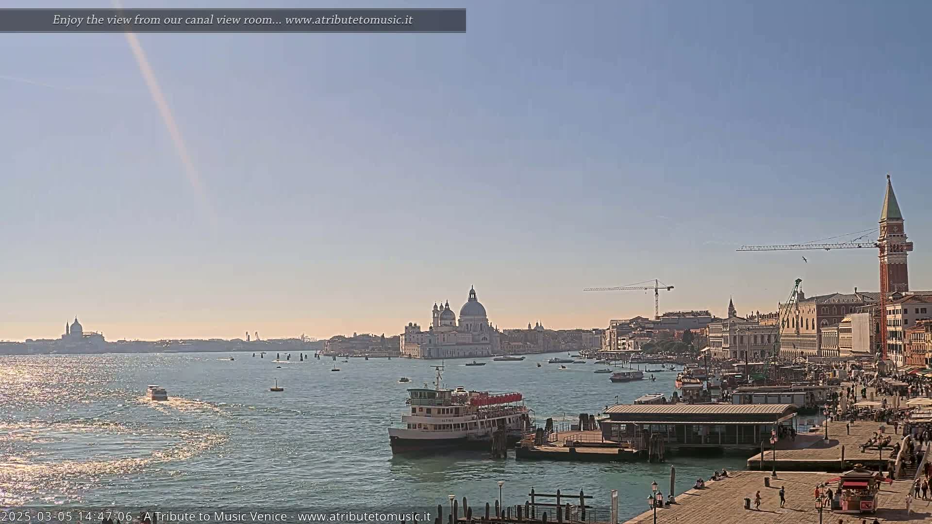 A sunny Venetian canal scene shows a large passenger ferry near a dock, with the Basilica di Santa Maria della Salute and San Giorgio Maggiore in the distance.