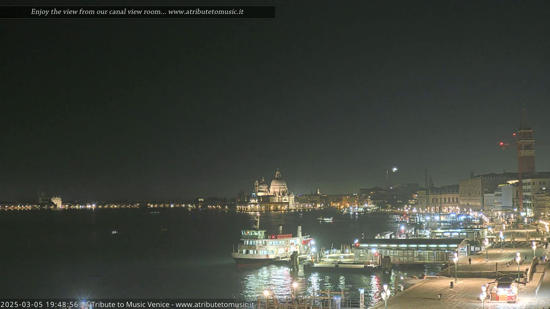 A nighttime view of a canal in Venice, Italy, shows a ferry docked near illuminated buildings and a church, under a clear, dark sky.