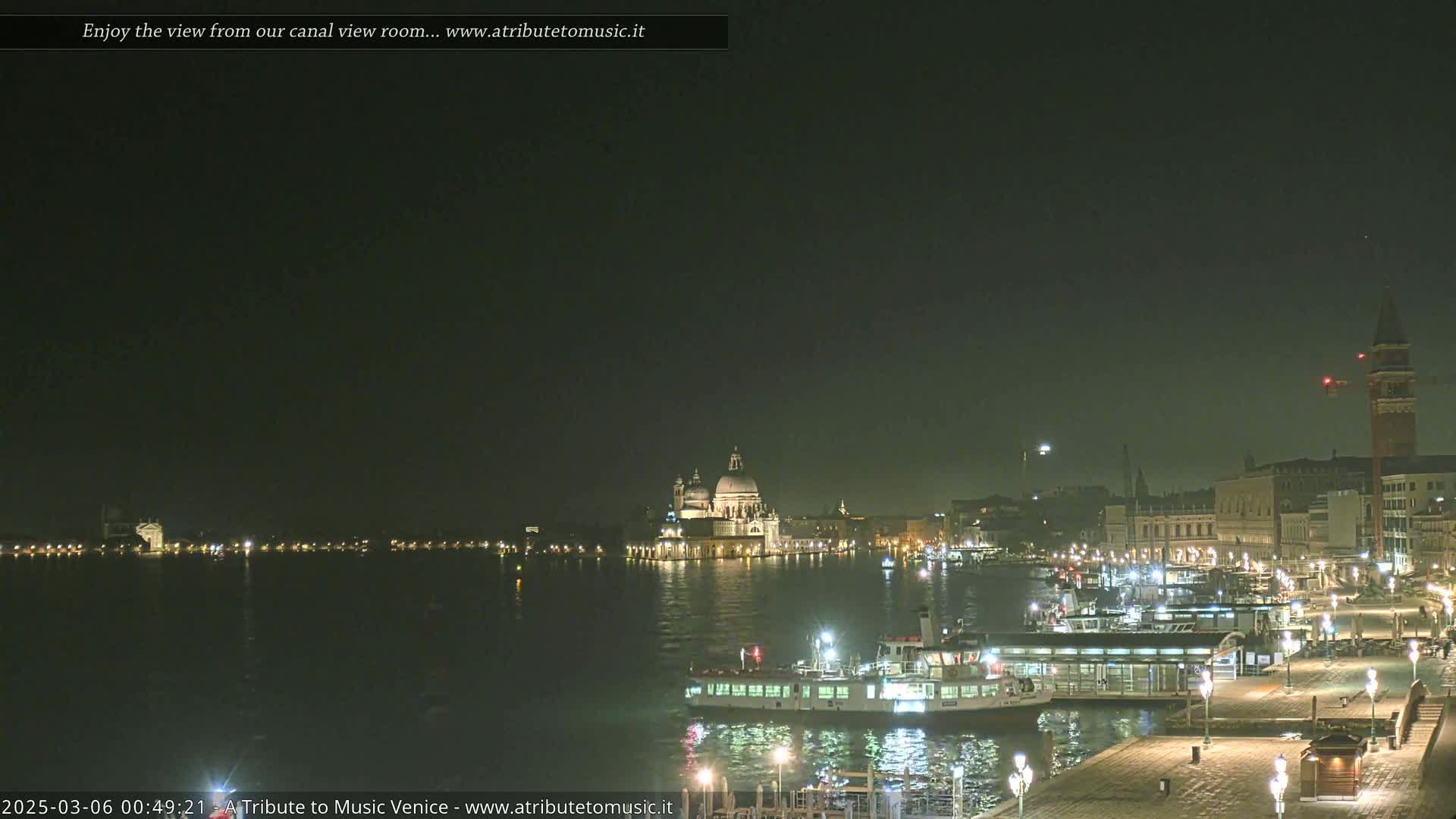A nighttime view of a canal in Venice, showing illuminated buildings, boats, and the Santa Maria della Salute church across the water under a dark, clear sky.