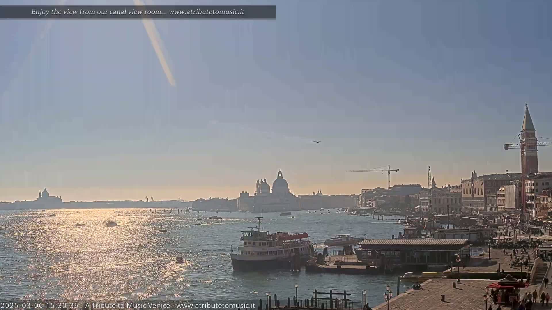 A sunny day shows a Venetian canal with a ferry, other boats, and buildings along the waterfront, including a prominent dome and tall bell tower in the distance.