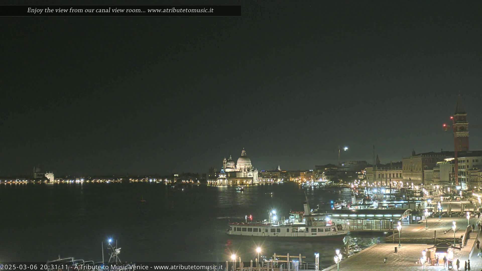A nighttime view across a canal shows illuminated buildings, boats, and a large church in Venice under a clear, dark sky.
