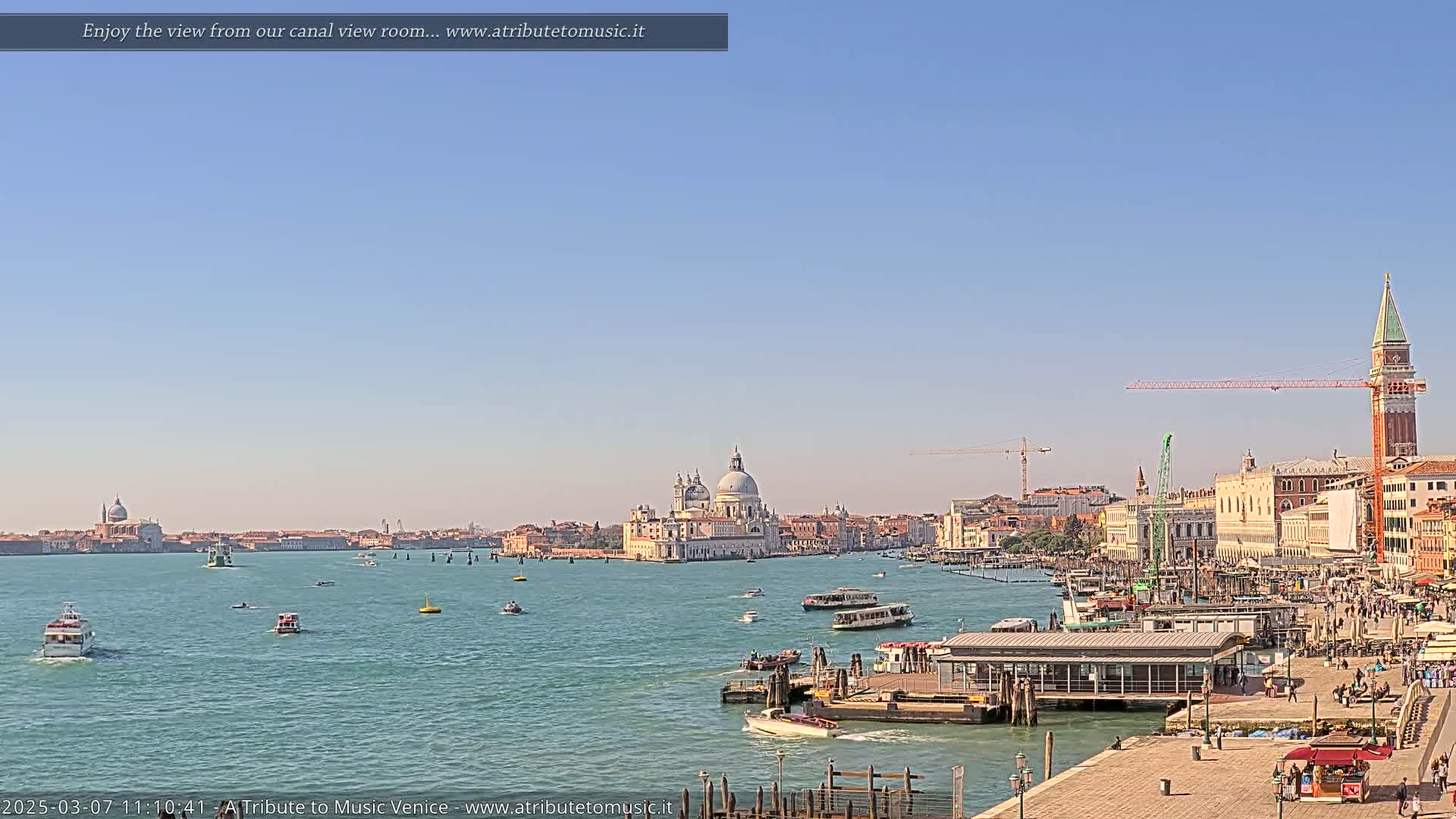 A sunny day in Venice shows a wide canal with boats, a cityscape featuring a large domed church, and a tall bell tower.