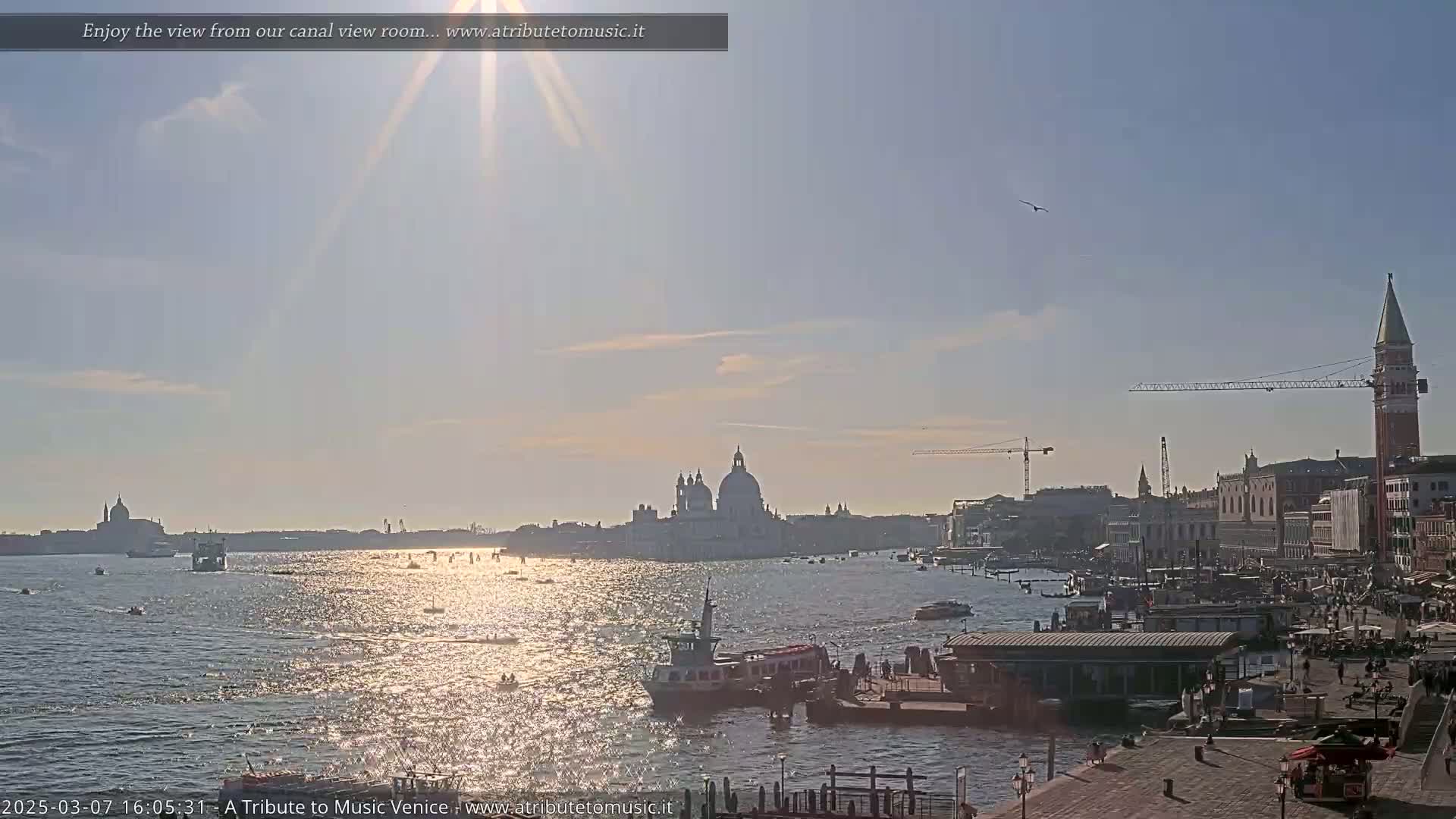 A sunny day in Venice shows a glittering canal with boats, a cityscape featuring a domed church and a tall bell tower, and construction cranes in the distance.
