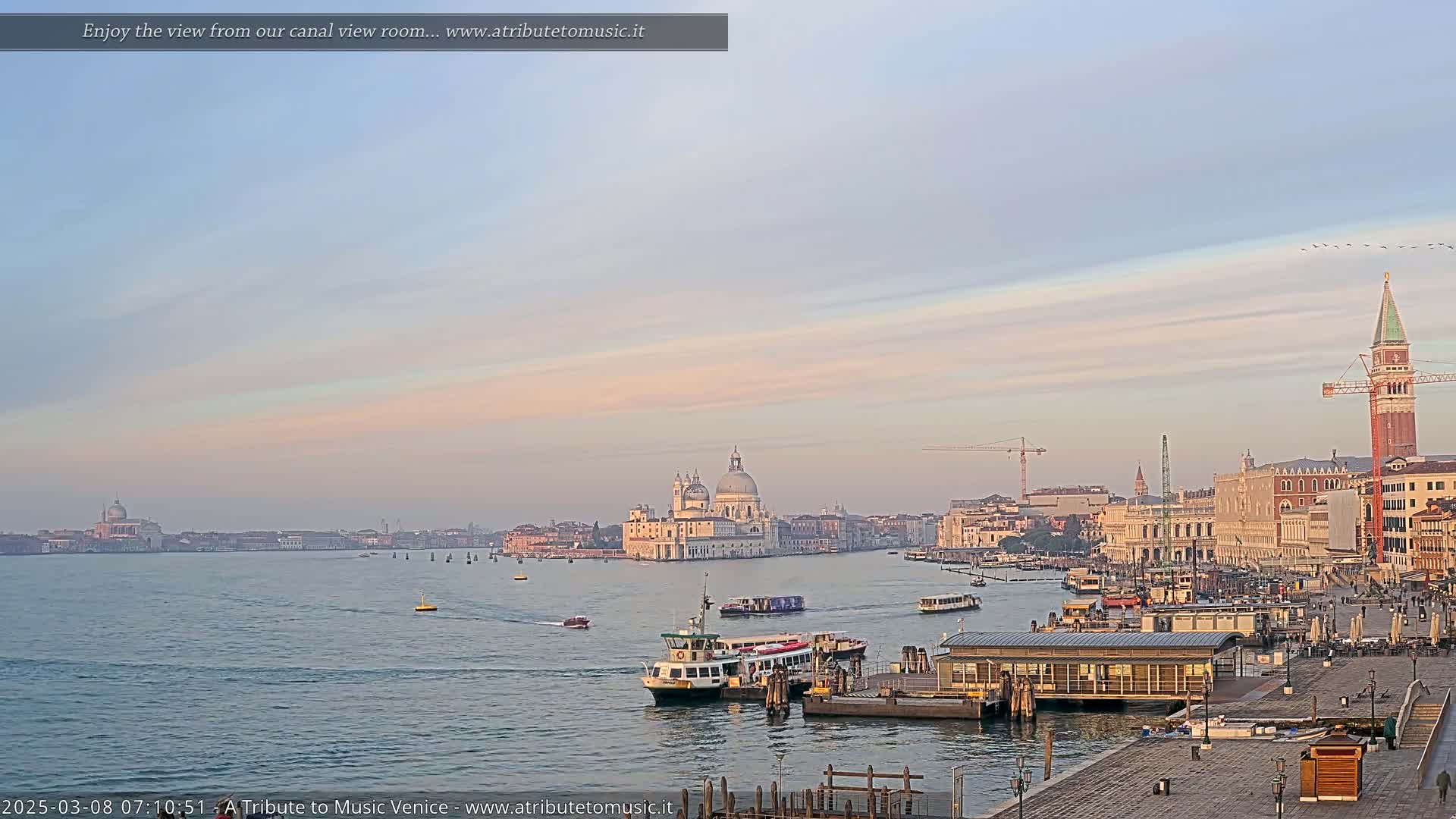 A calm waterway shows a Venetian cityscape under a pale sunrise sky, with boats on the water and buildings including a large domed church in the background.
