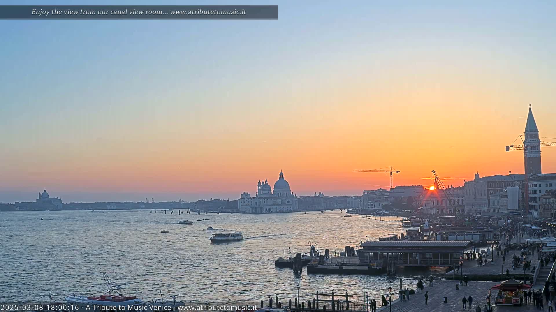 A sunset over a Venetian canal shows boats on the water, buildings along the shore, and a large church in the distance.