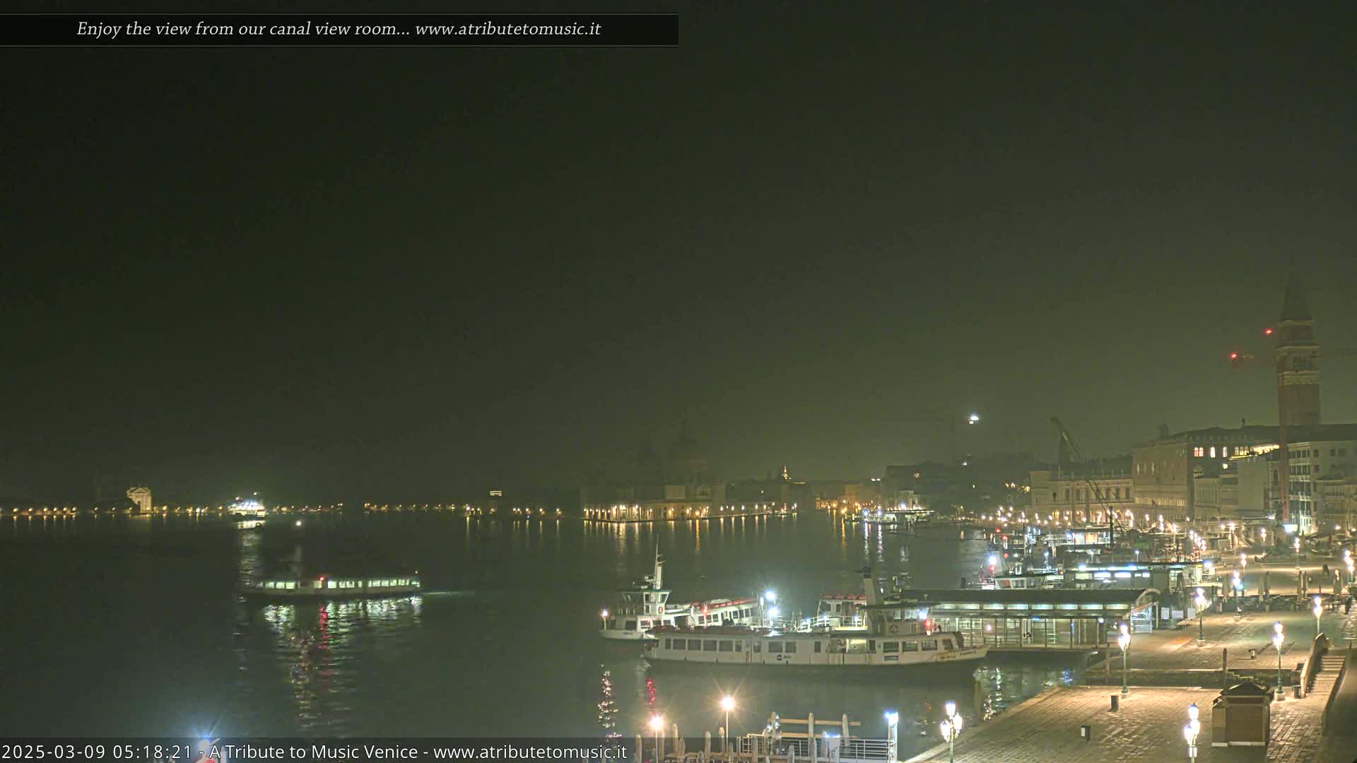 A nighttime view of a canal in Venice, Italy, shows several boats moored near a waterfront, with buildings and lights visible in the hazy distance.