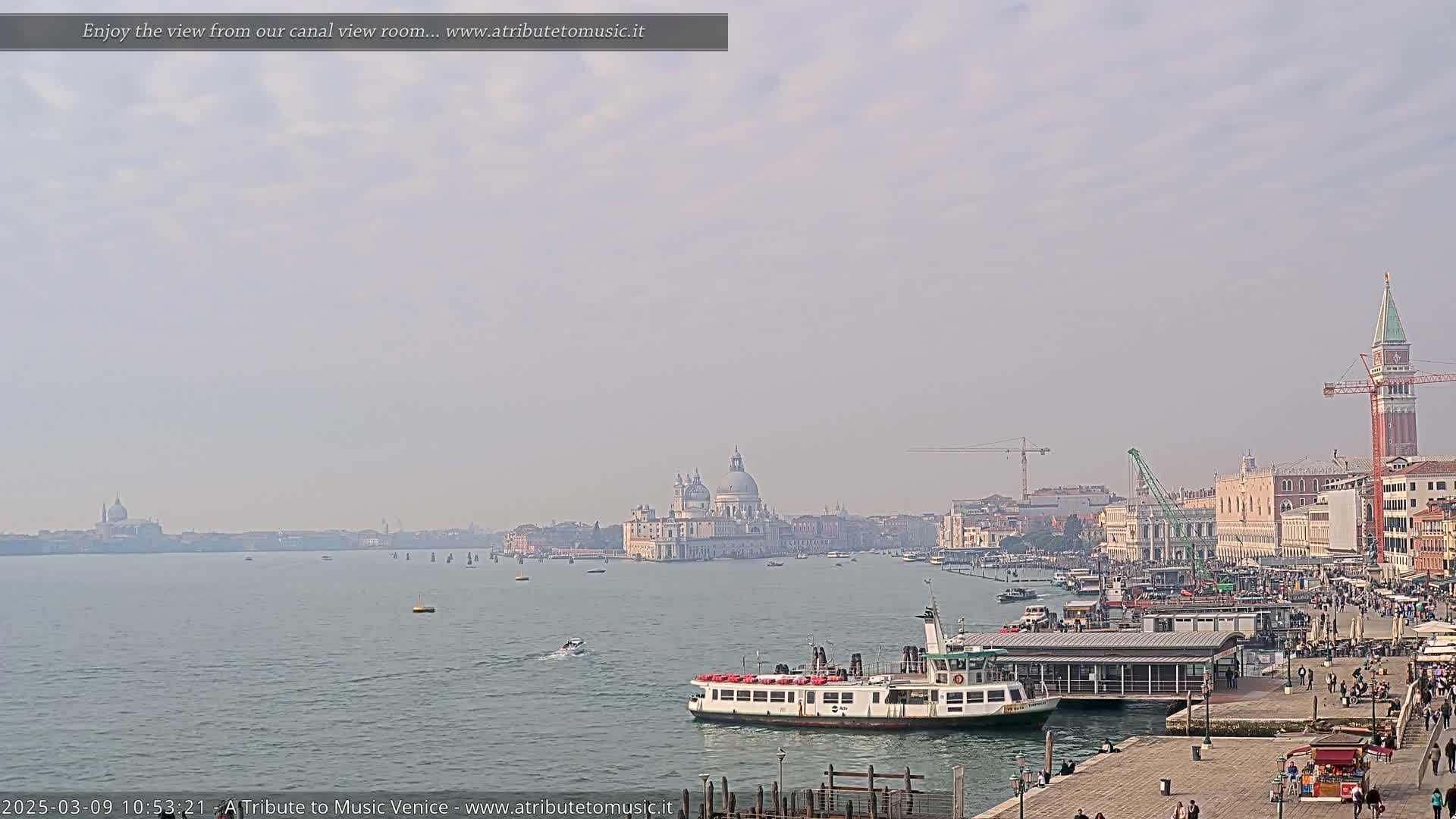 A hazy day in Venice shows a large body of water with a ferry and several other small boats, with the Basilica di Santa Maria della Salute and other buildings visible in the distance.