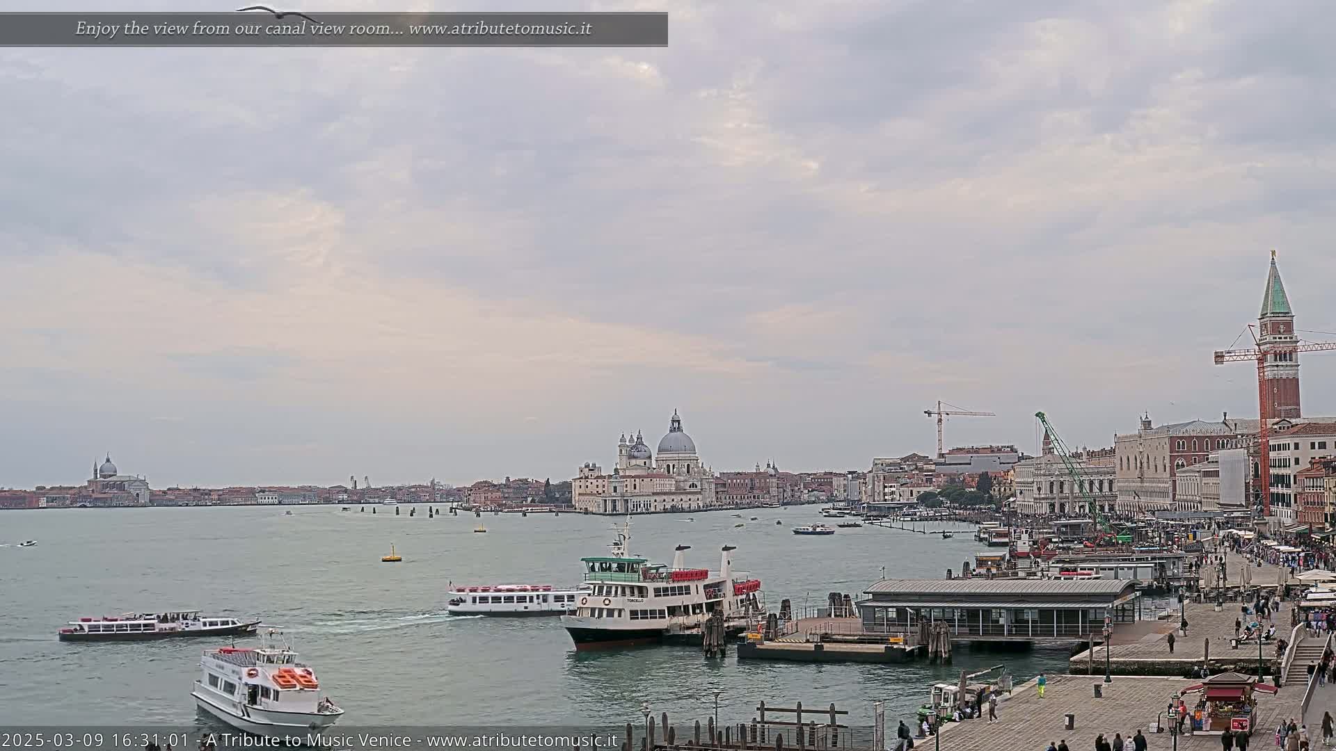 Several water taxis and passenger ferries navigate a wide canal in Venice under a cloudy sky, with the Basilica di Santa Maria della Salute and other buildings visible in the distance.