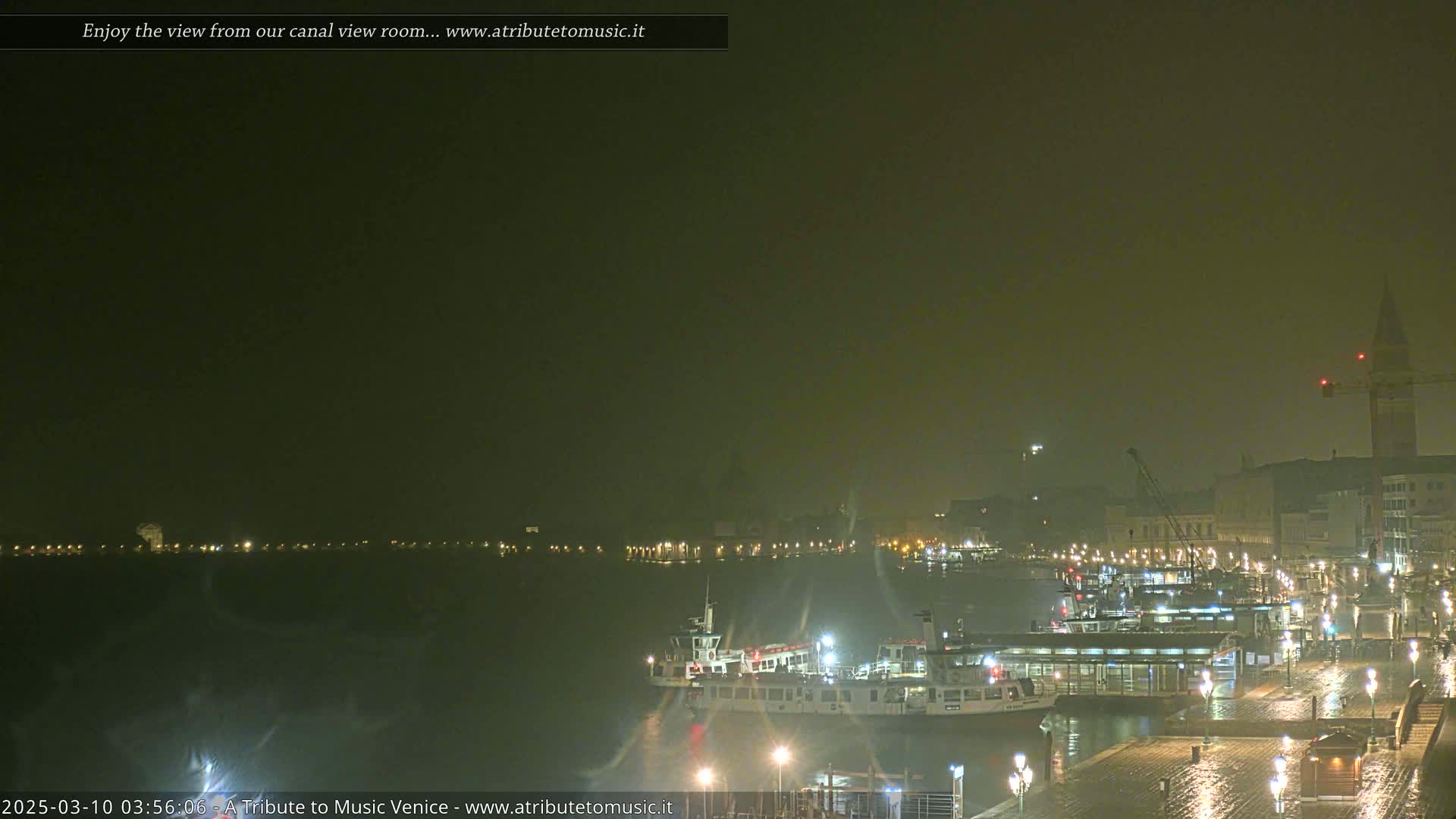 A nighttime view across a canal shows a hazy cityscape with several boats docked, lit by streetlights and the boats' own lights, in rainy weather.
