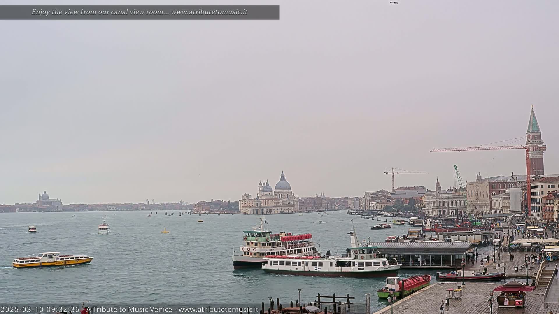 A view across a calm canal in Venice, showing several water buses and the city skyline under an overcast sky.