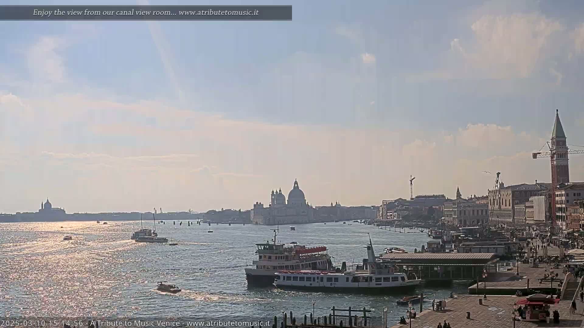 A sunny day in Venice shows a wide canal with several boats, buildings lining the far shore, and the bell tower of San Giorgio Maggiore in the distance.