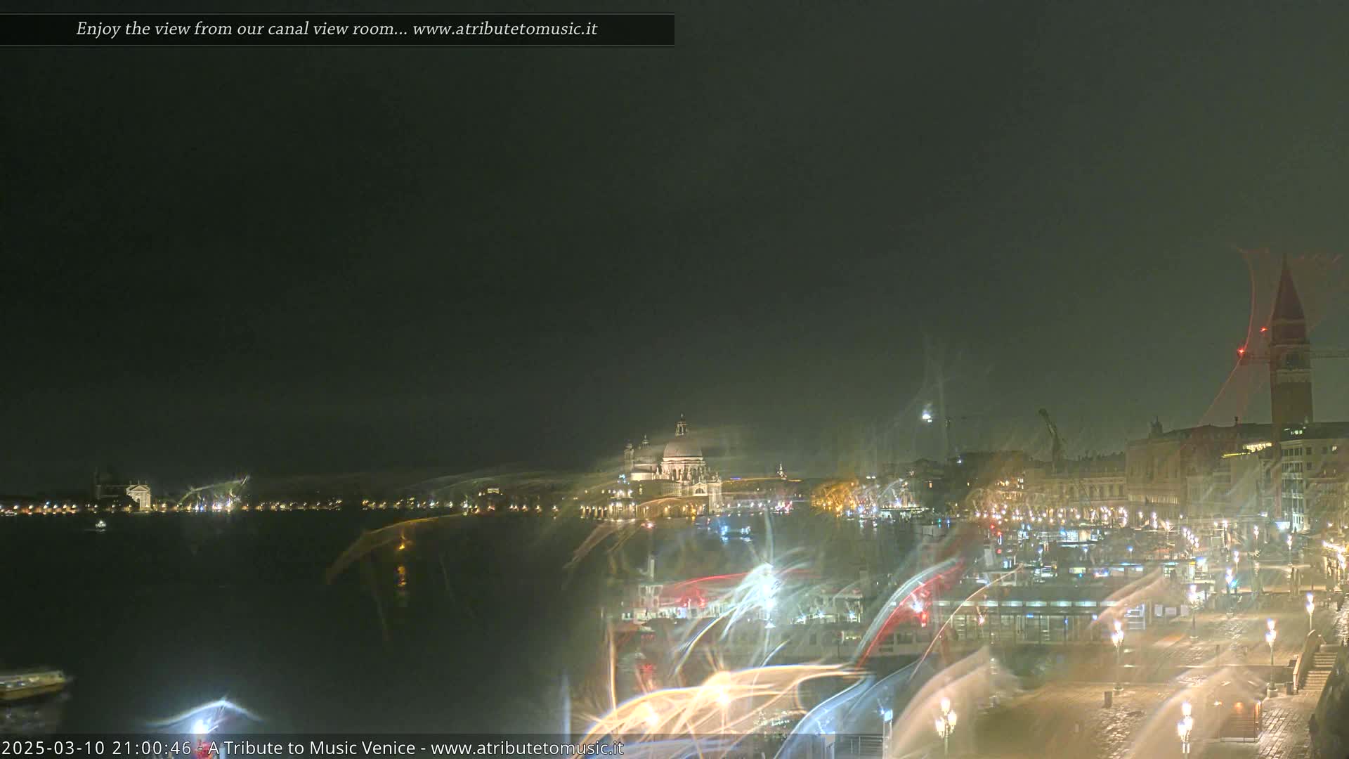 A nighttime view of a canal city, possibly Venice, shows illuminated buildings and a large dome-shaped structure across the water in hazy or foggy conditions.