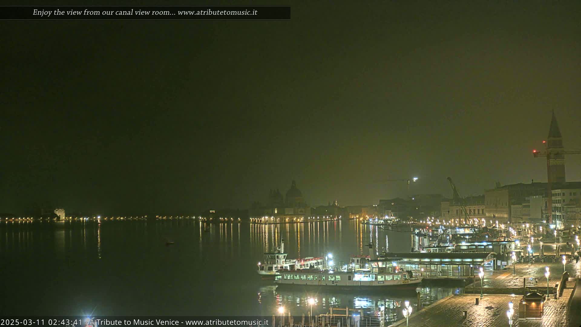 A nighttime view of a canal in Venice, Italy, shows several boats docked near illuminated buildings under a hazy, dark sky.