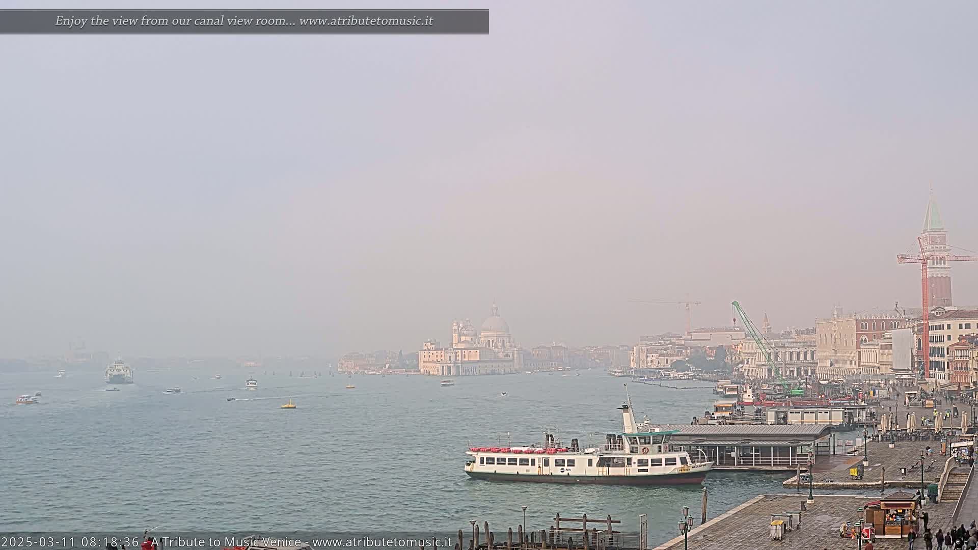 A hazy view of a Venetian canal shows several boats, a ferry at a dock, and buildings including a large church in the distance.