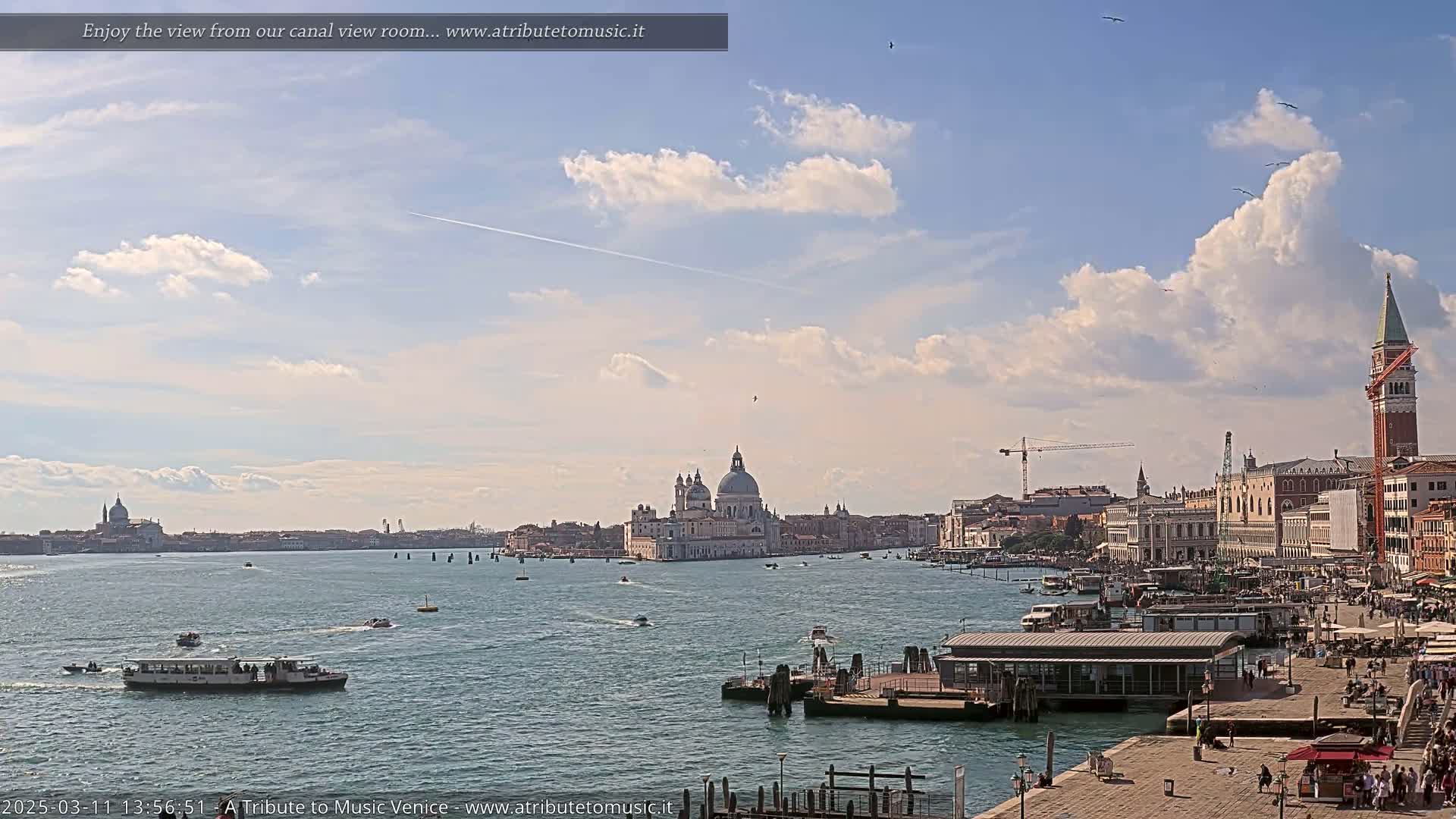 A wide shot of a Venetian waterway on a partly cloudy day shows boats on the water, buildings lining the shore, and a large domed church in the distance.