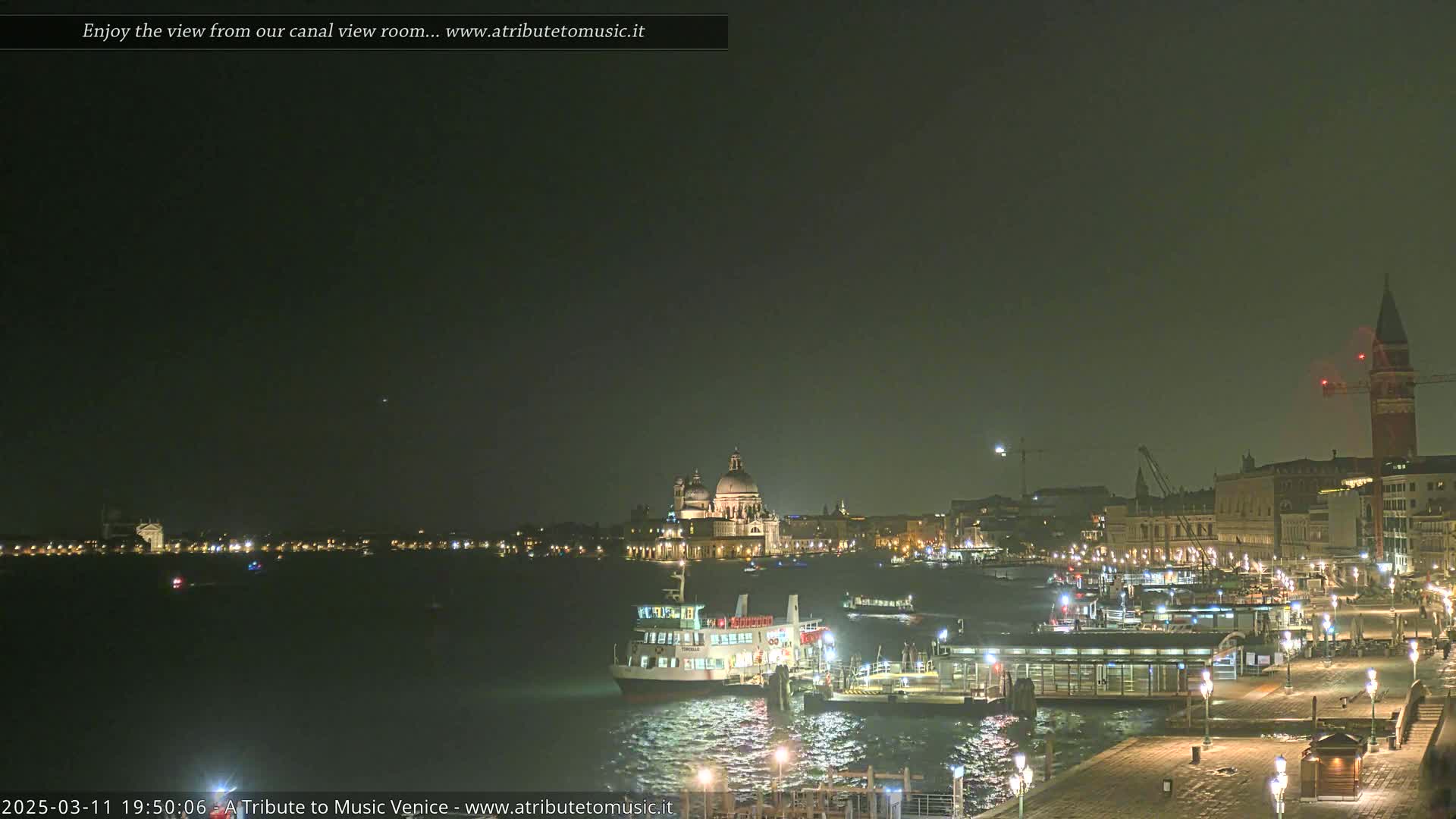 A nighttime view of a Venetian canal, with boats docked at a waterfront station, and the Basilica di Santa Maria della Salute illuminated in the distance under a dark, clear sky.