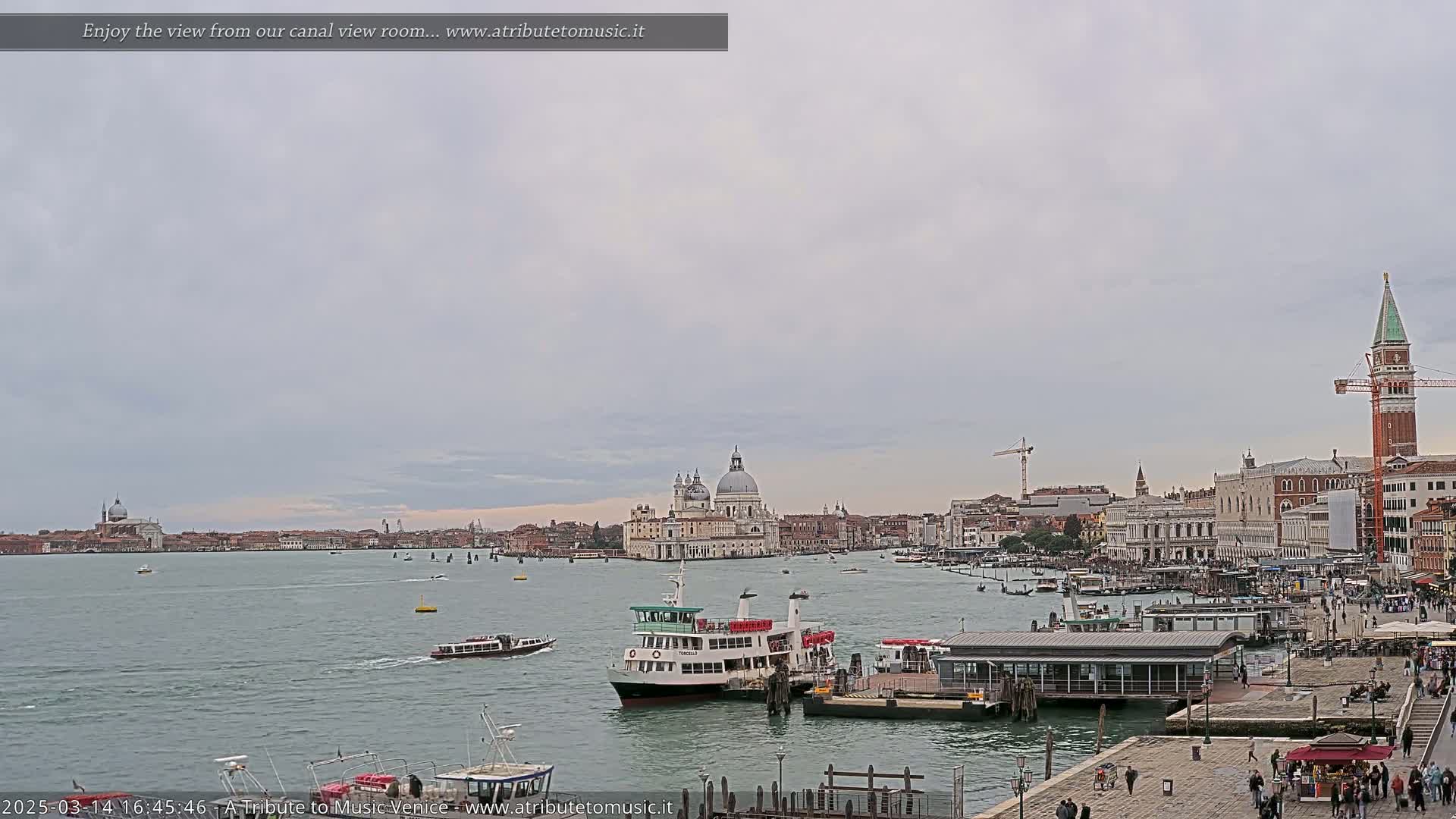 A wide, calm canal with several boats, including a large passenger ferry, under an overcast sky, showing the city of Venice in the background.