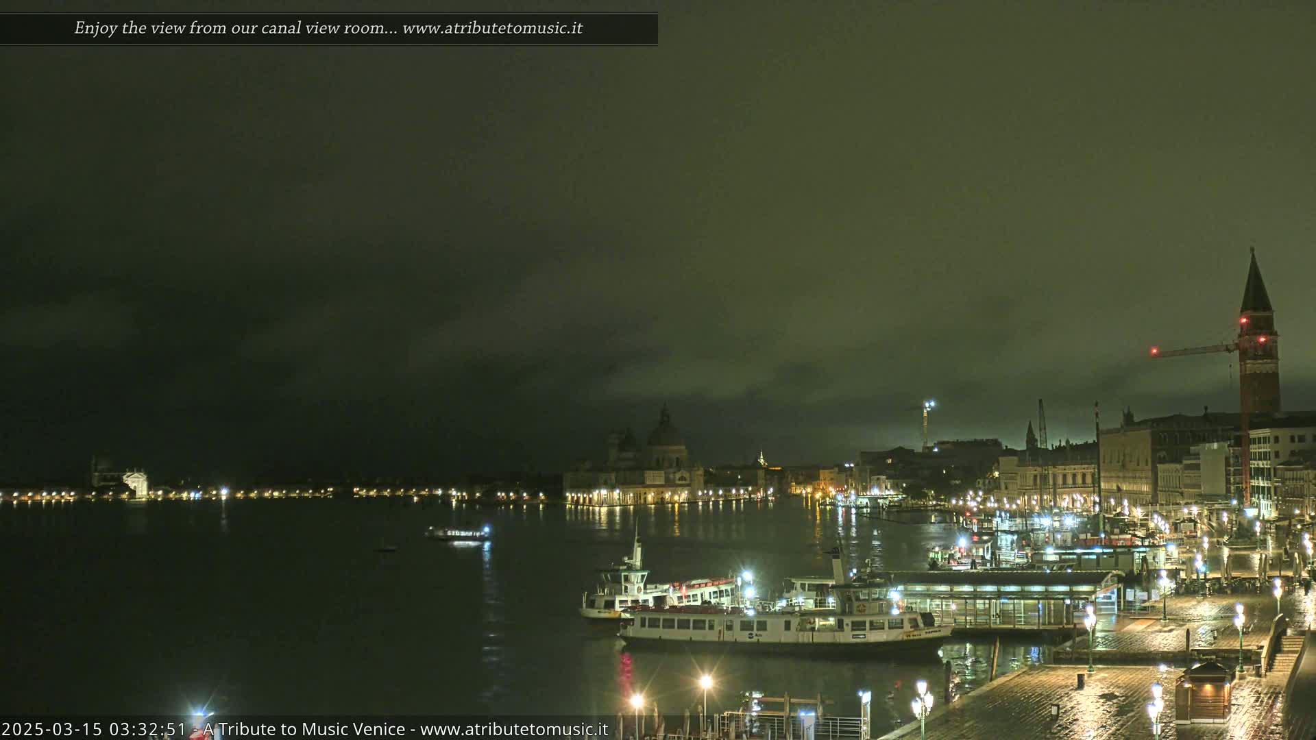 A nighttime view of a canal in Venice, Italy, shows several boats docked along a waterfront under a dark, cloudy sky.