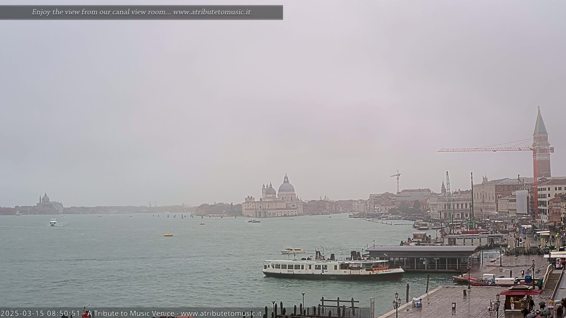 A hazy, overcast view of a canal in Venice, showing boats, buildings, and a large church in the distance.