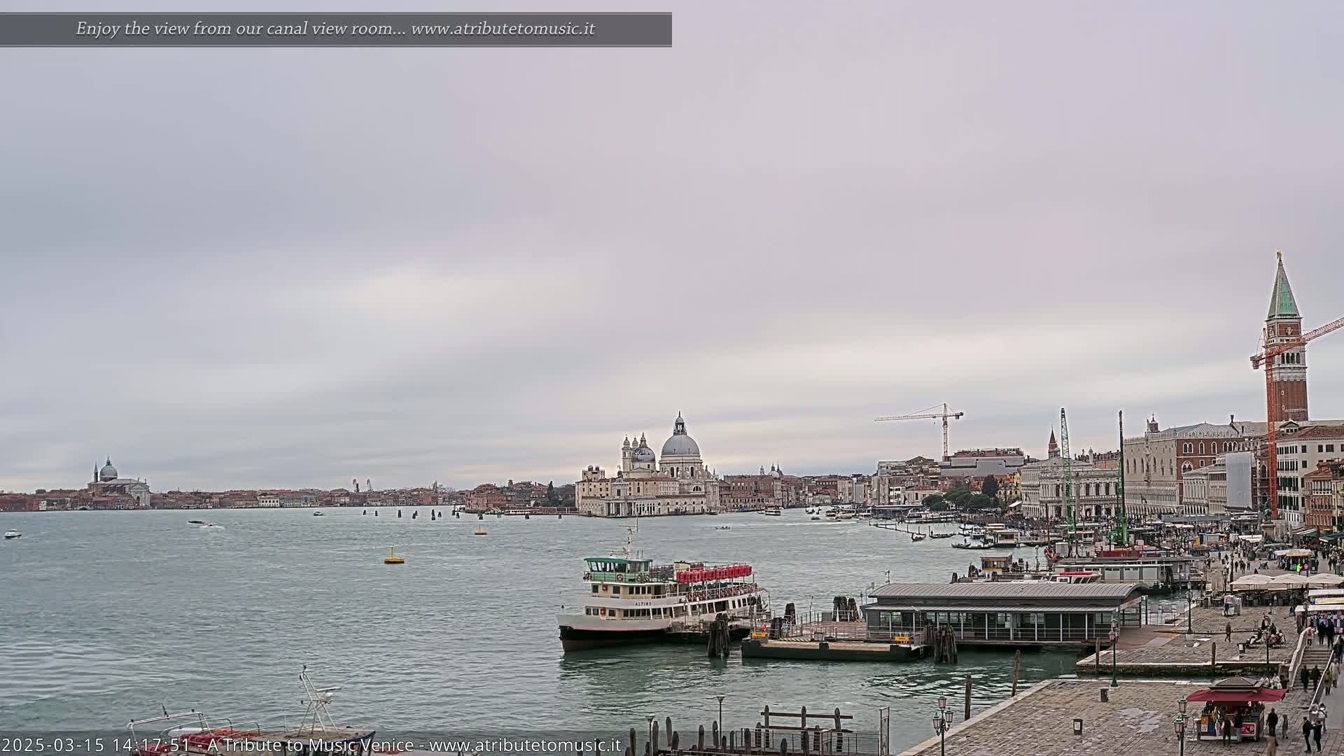 A ferry is docked near a waterfront in Venice, Italy, under an overcast sky, with the Basilica di Santa Maria della Salute visible in the distance.