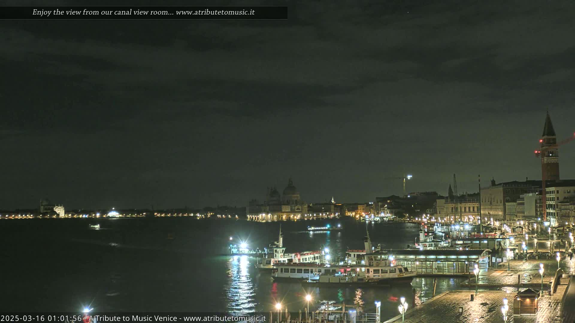 A nighttime view of a waterway with numerous illuminated boats and buildings along the shore under a dark, cloudy sky.