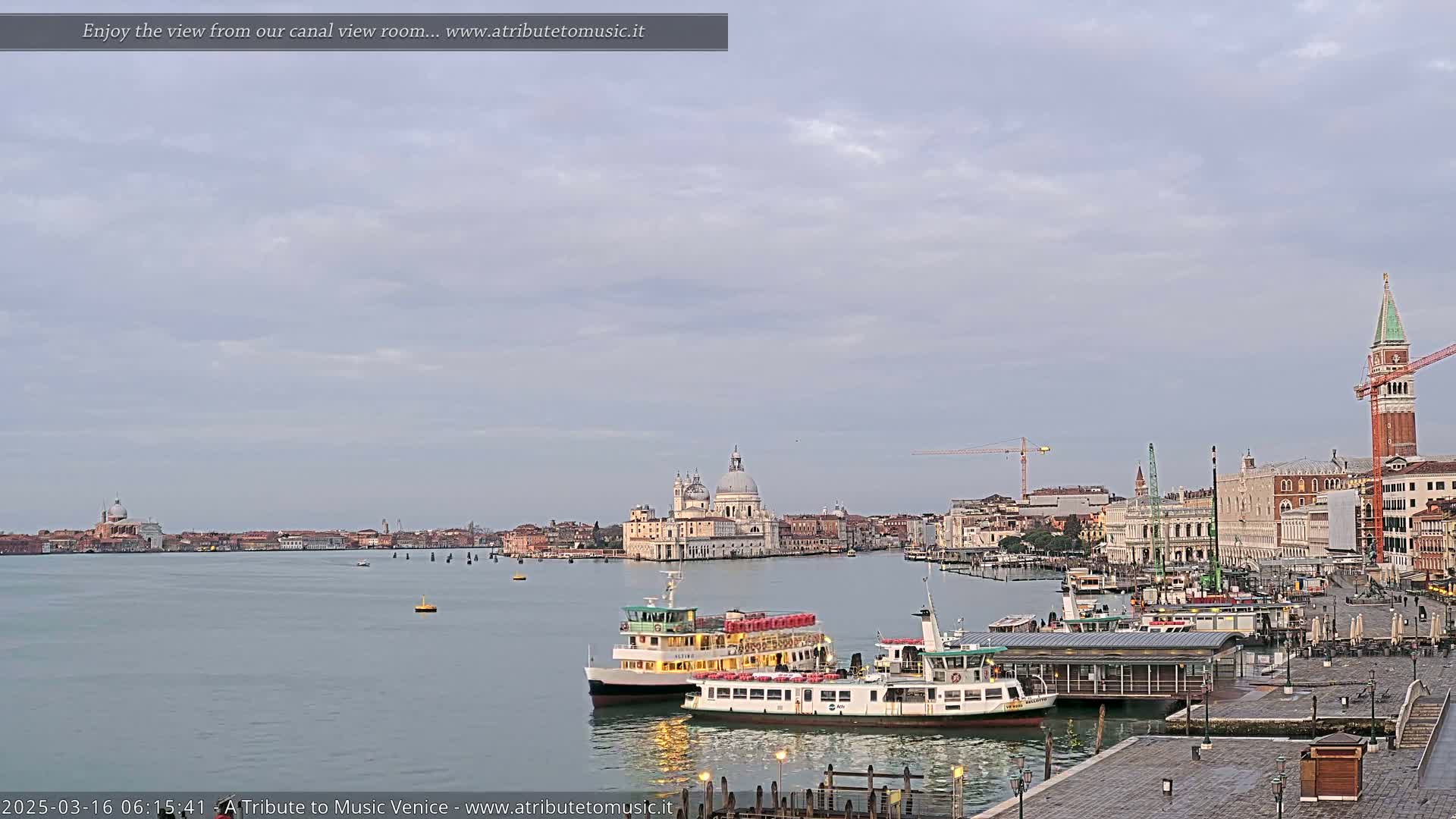 A calm canal scene in Venice shows two ferries docked near a waterfront plaza, with the Basilica di Santa Maria della Salute and St. Mark's Campanile visible in the distance under a cloudy sky.