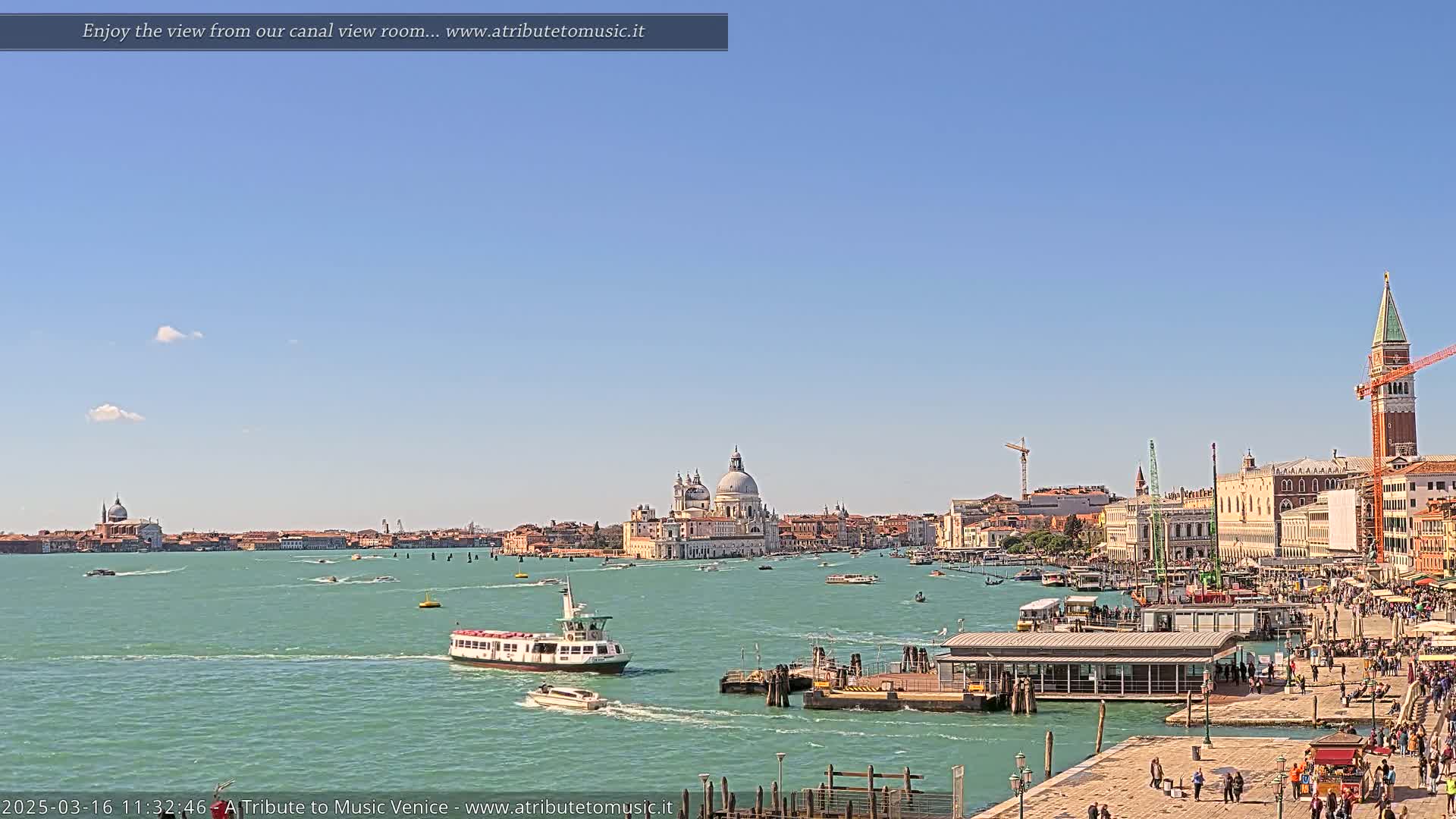 A sunny day in Venice shows a waterway with boats, a ferry, and a view of the city skyline, including a large domed church and a bell tower.