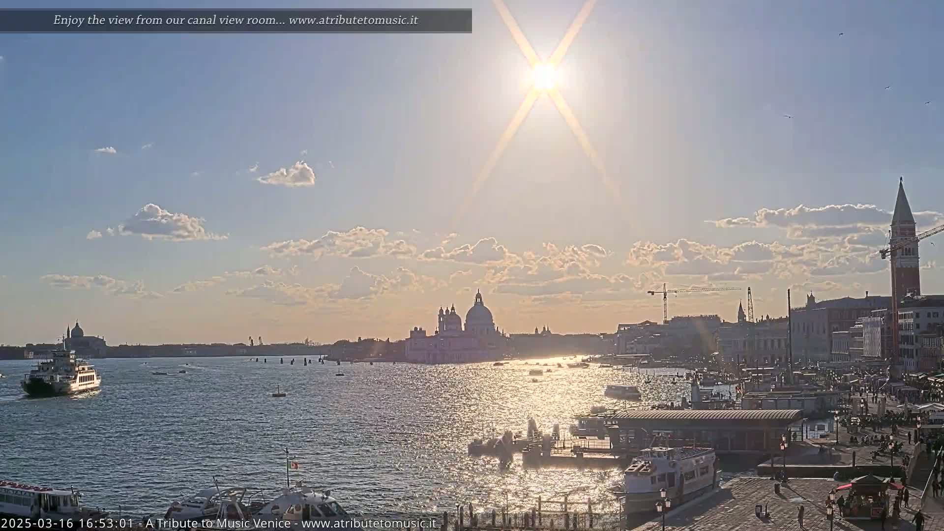A sunny day in Venice shows a wide canal with boats, buildings along the waterfront, and the Basilica di Santa Maria della Salute in the distance.