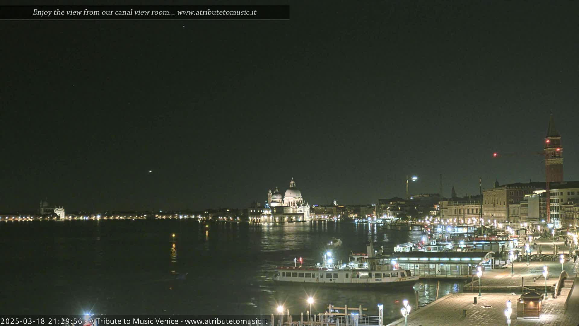 A nighttime view of a canal in Venice, Italy, shows illuminated buildings, boats, and a clear, dark sky.